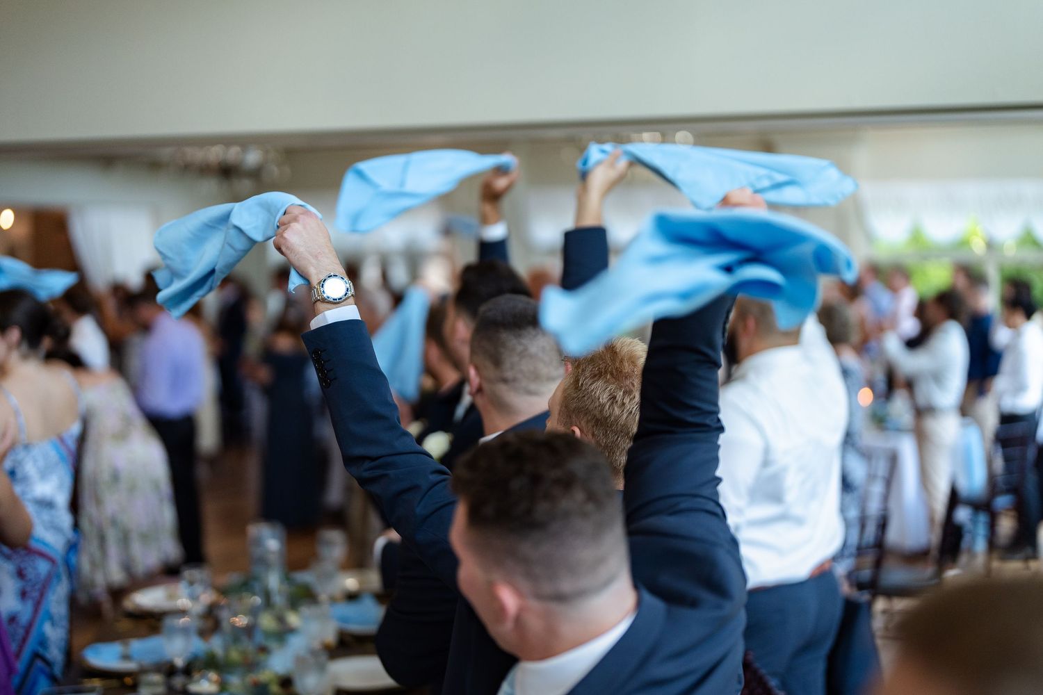 Wedding guests wave blue napkins in celebration during lively indoor wedding reception dinner party.