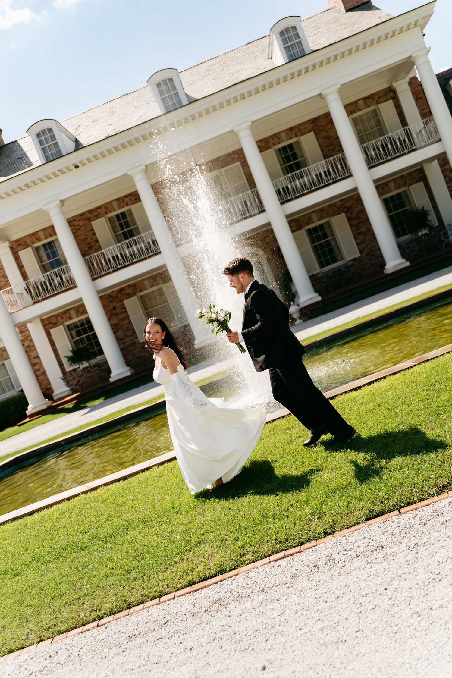 A couple in wedding attire runs playfully on the lawn in front of a grand white colonial mansion with columns.