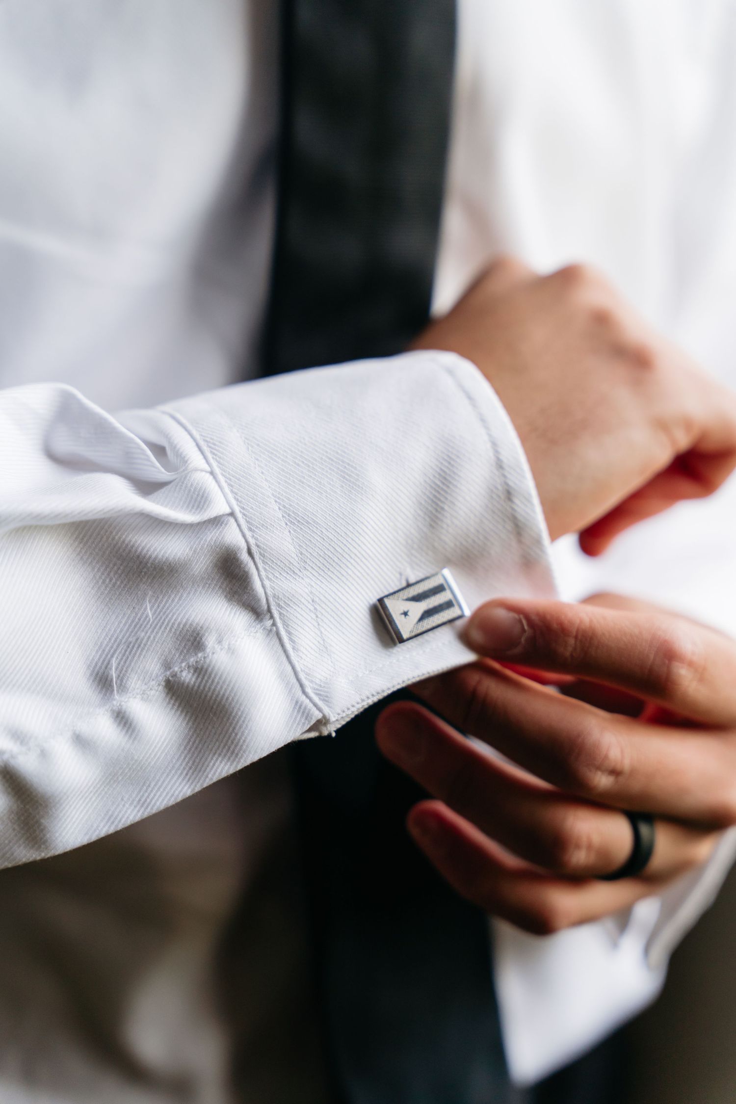 Close-up of hands adjusting a silver cufflink on a white dress shirt sleeve with a dark tie visible in background.