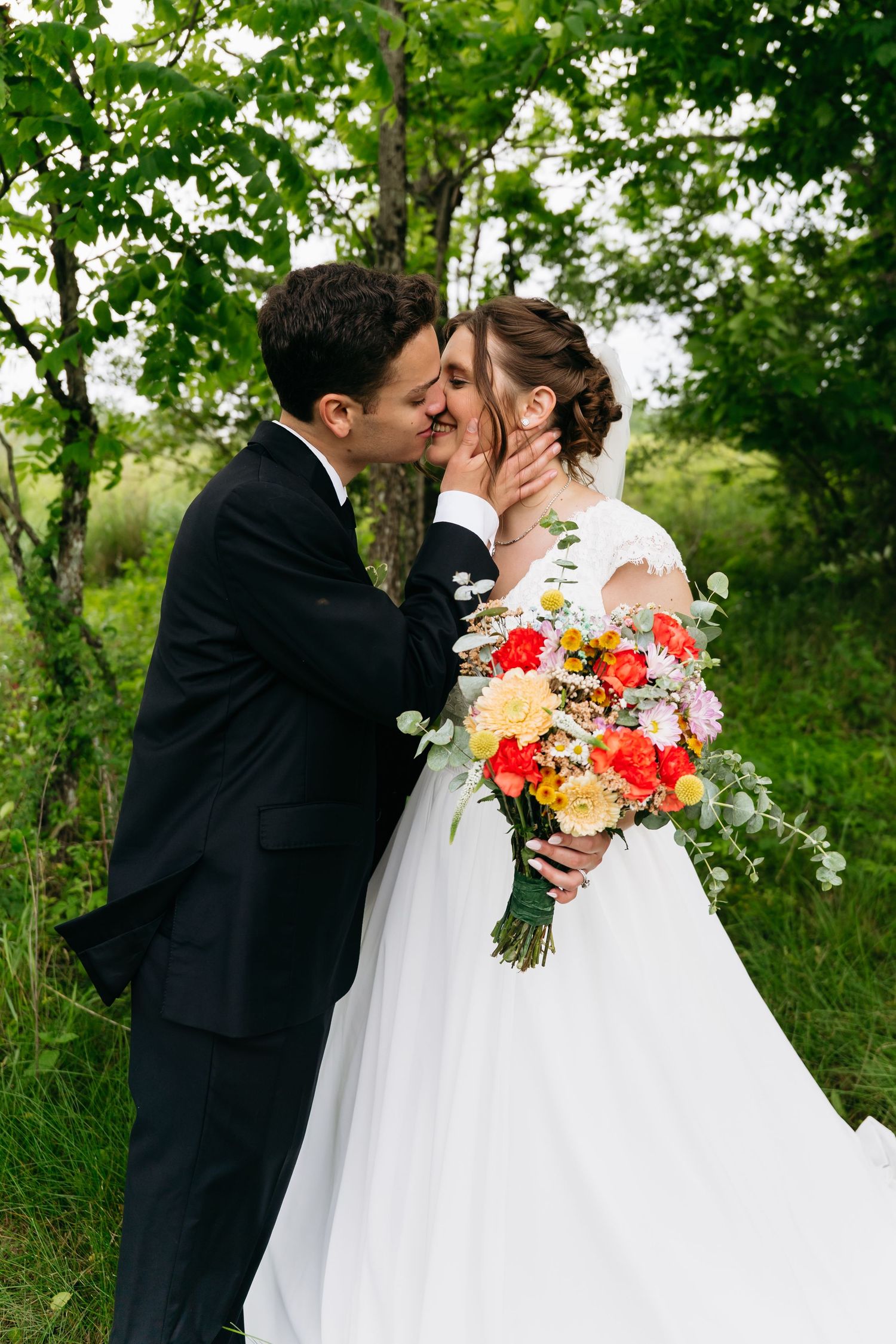 Newlywed couple shares a romantic kiss outdoors surrounded by lush green foliage while holding a vibrant floral bouquet.