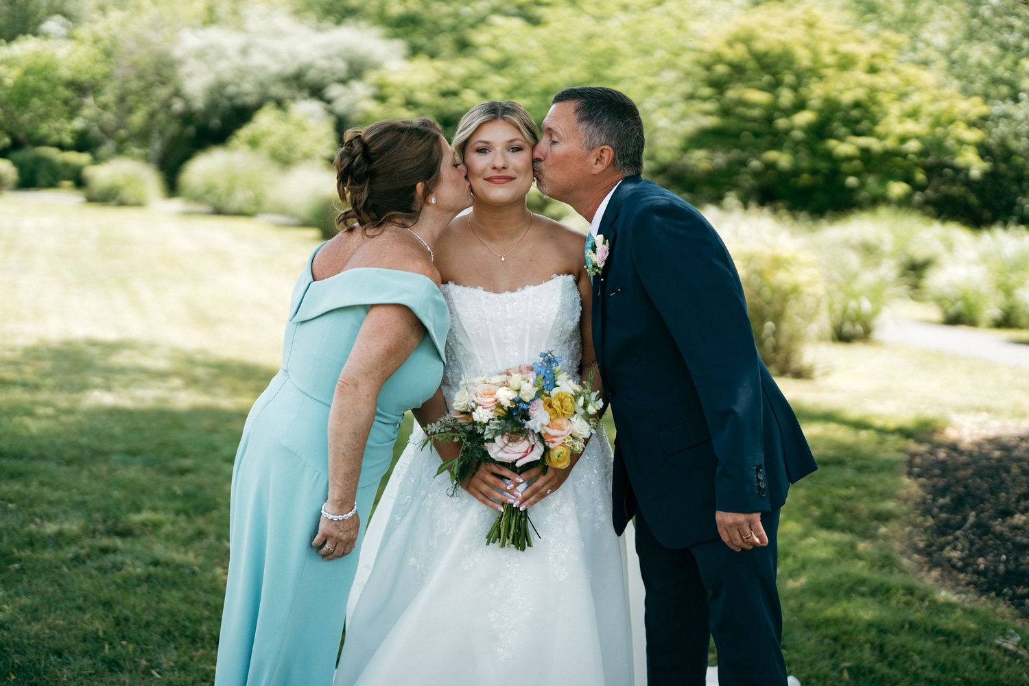 Bride holding colorful bouquet receives kisses from parents in blue attire on lush green garden wedding day.