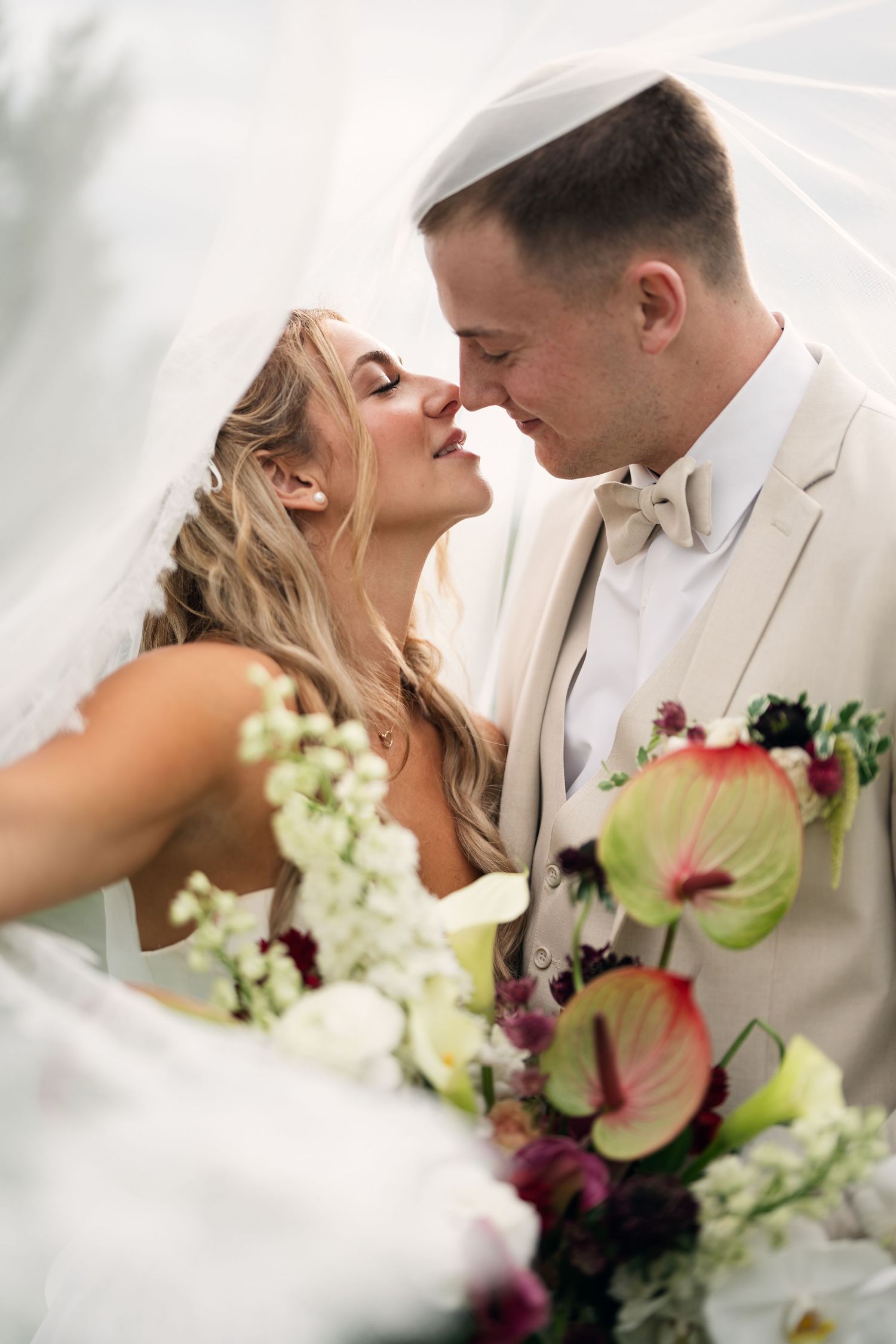Bride and groom share a romantic almost-kiss under a veil, surrounded by colorful floral bouquets at their wedding.