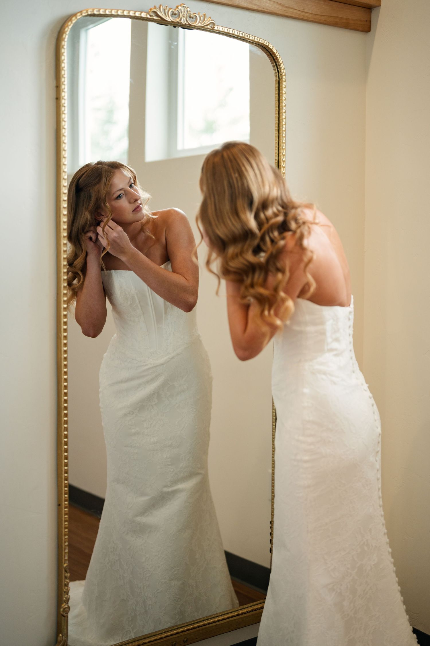 Bride in strapless lace wedding gown adjusting earring while gazing into ornate gold floor mirror.