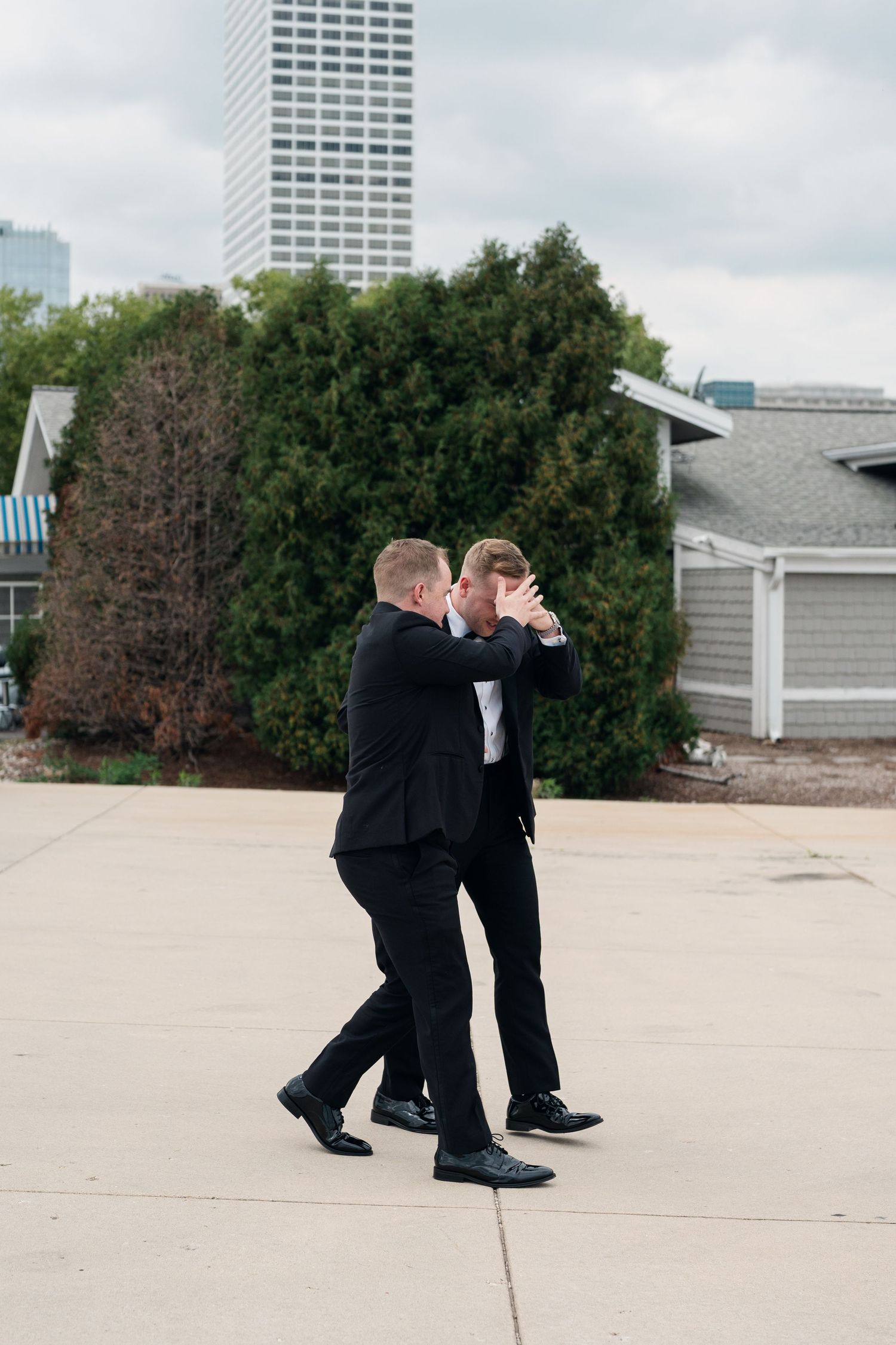 Two men in black suits share an emotional moment outdoors with city buildings and greenery in the background.