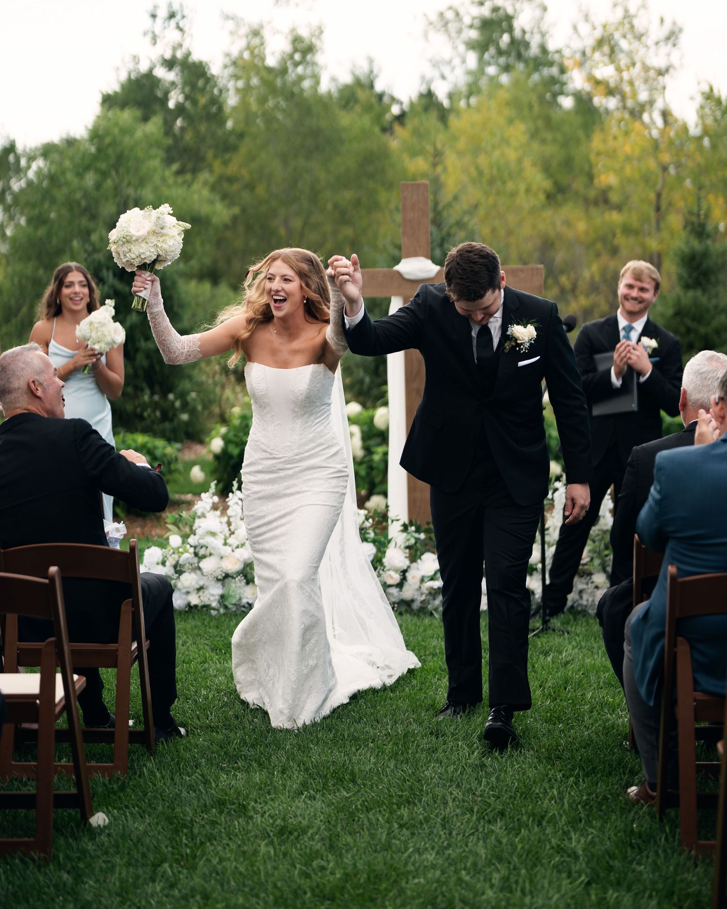 Joyful newlyweds walking back down the aisle after outdoor wedding ceremony, bride in white gown holding bouquet.