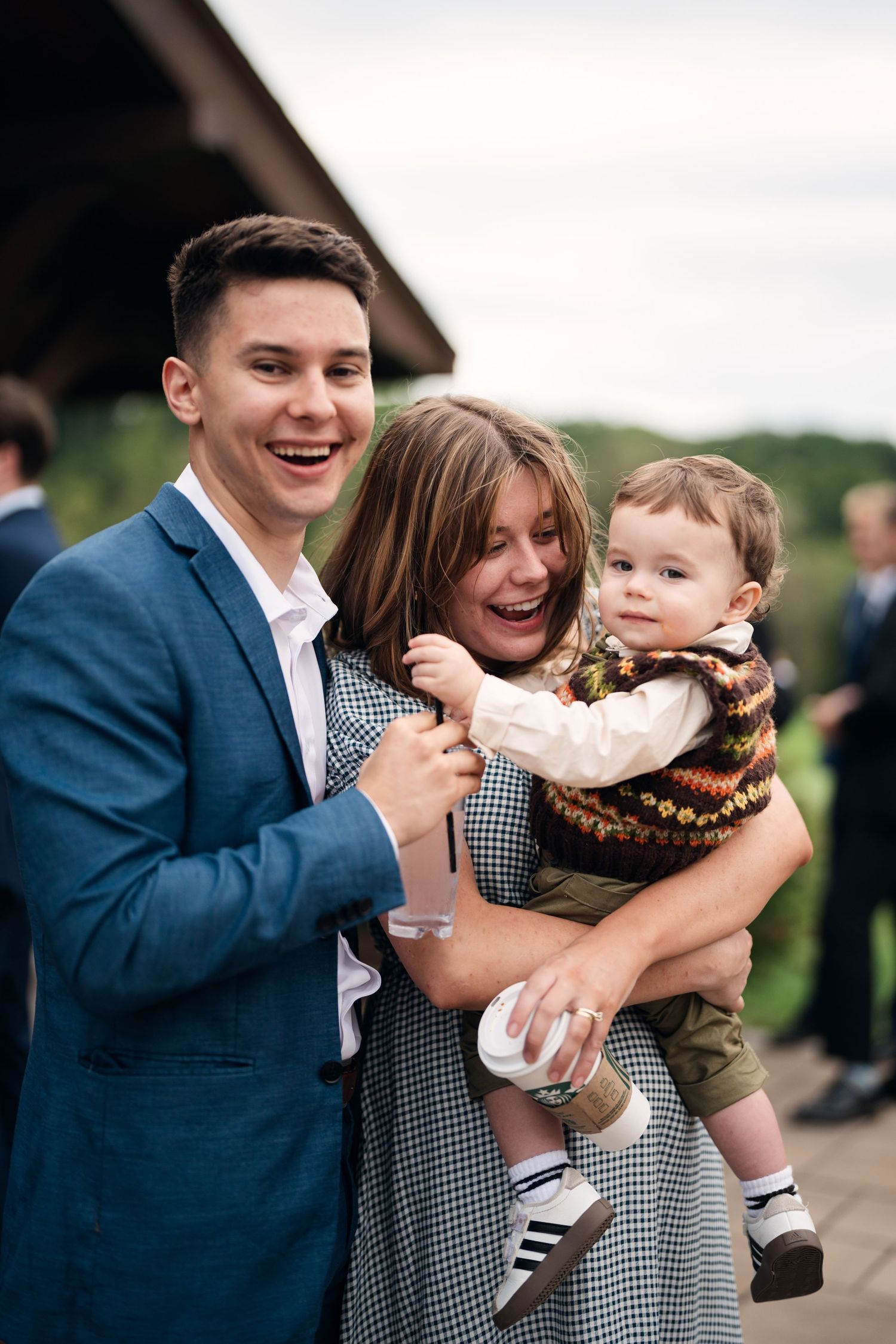 A laughing young man in a blue suit and woman joyfully hold a curious baby boy at an outdoor wedding reception.