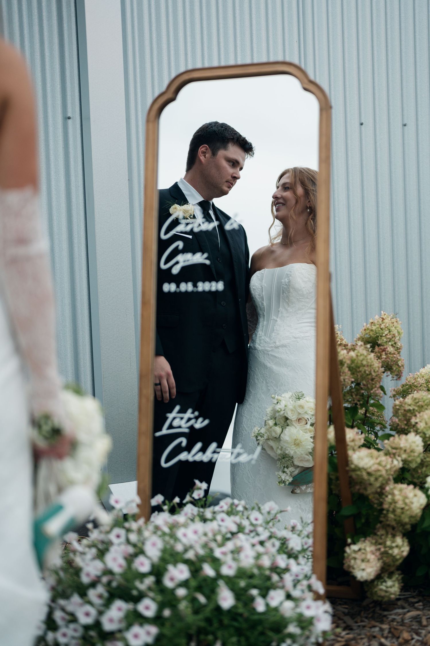 Couple reflected in gold-framed mirror with wedding date signage surrounded by floral arrangements.