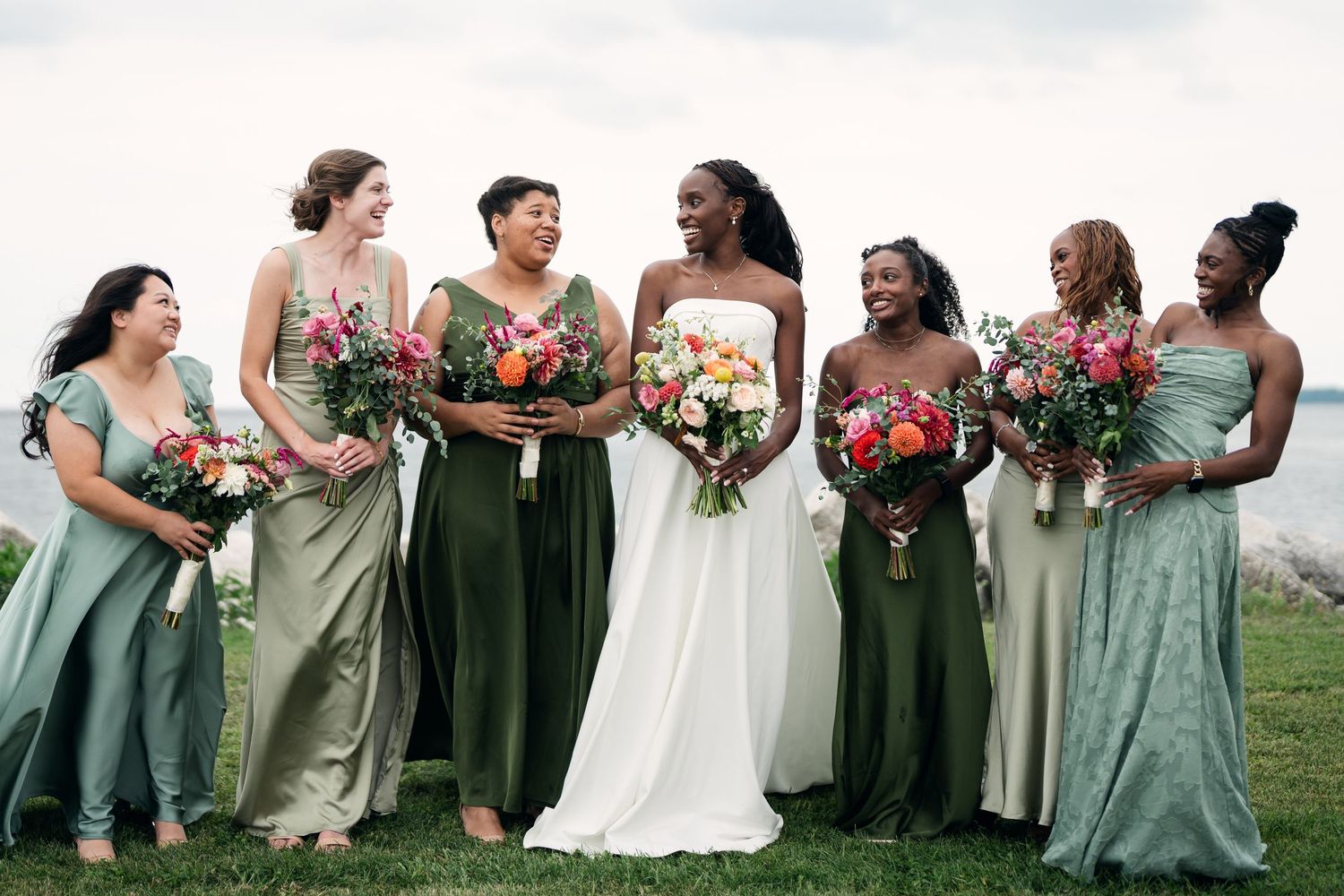 Bride in white gown with bridesmaids in mixed green dresses holding colorful bouquets outdoors near water.