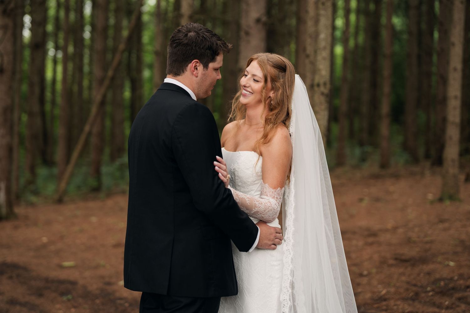 Bride and groom share a tender moment in a forest, holding hands as she smiles up at him in her white gown and veil.