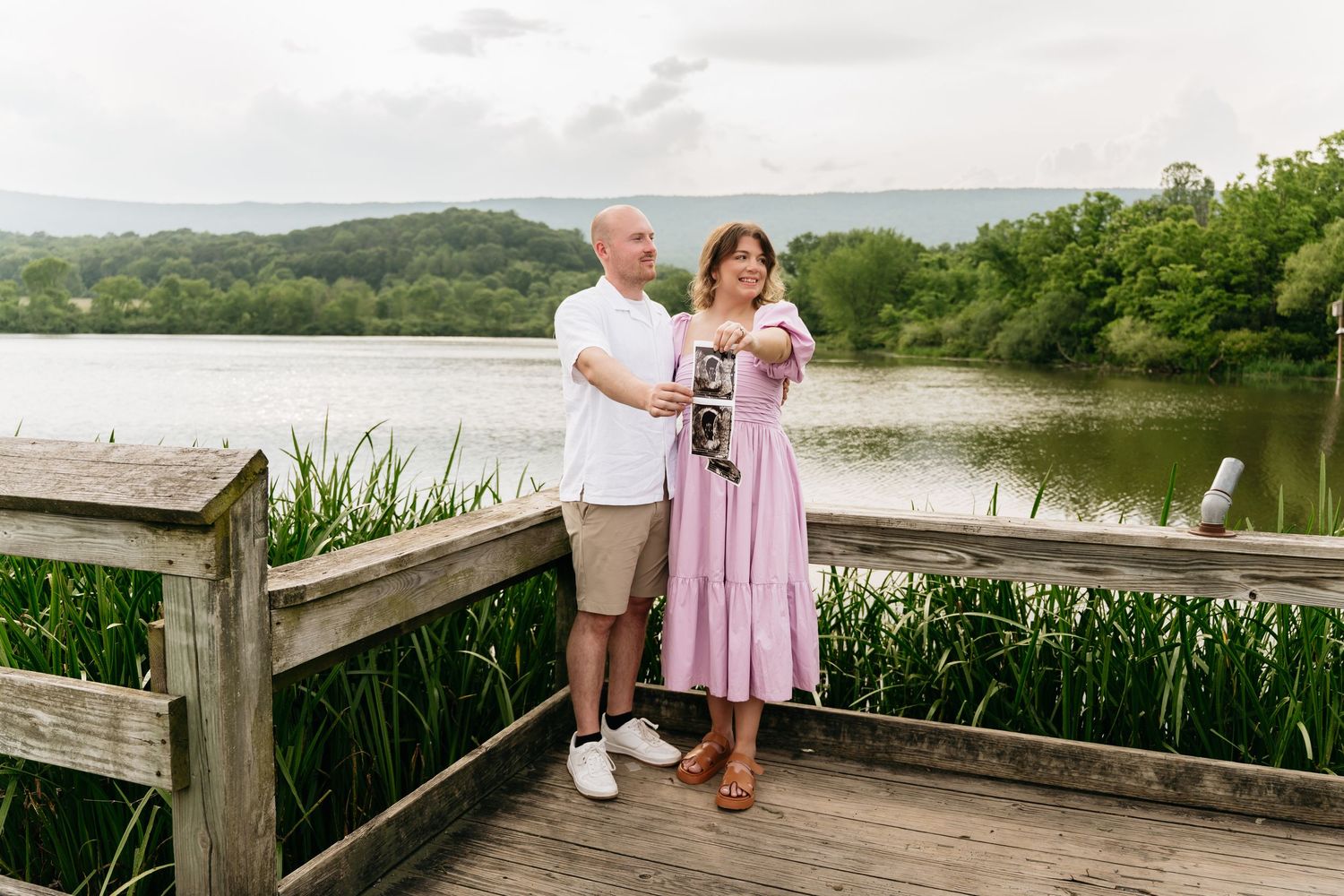 A couple sits on a rustic wooden boardwalk overlooking a scenic coastal marsh during golden hour.