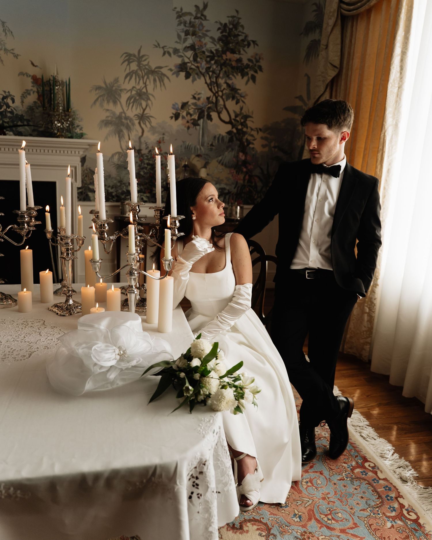 Elegant couple poses by candlelit wedding table with ornate decorations and flowing white gown in vintage-styled room.