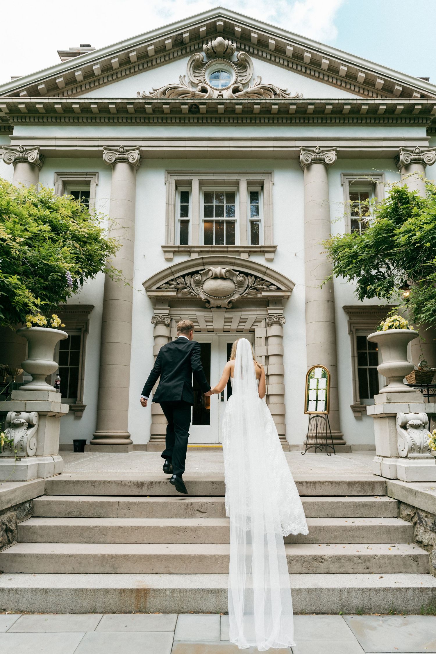 Bride and groom walking hand-in-hand up stone steps toward grand neoclassical wedding venue entrance.