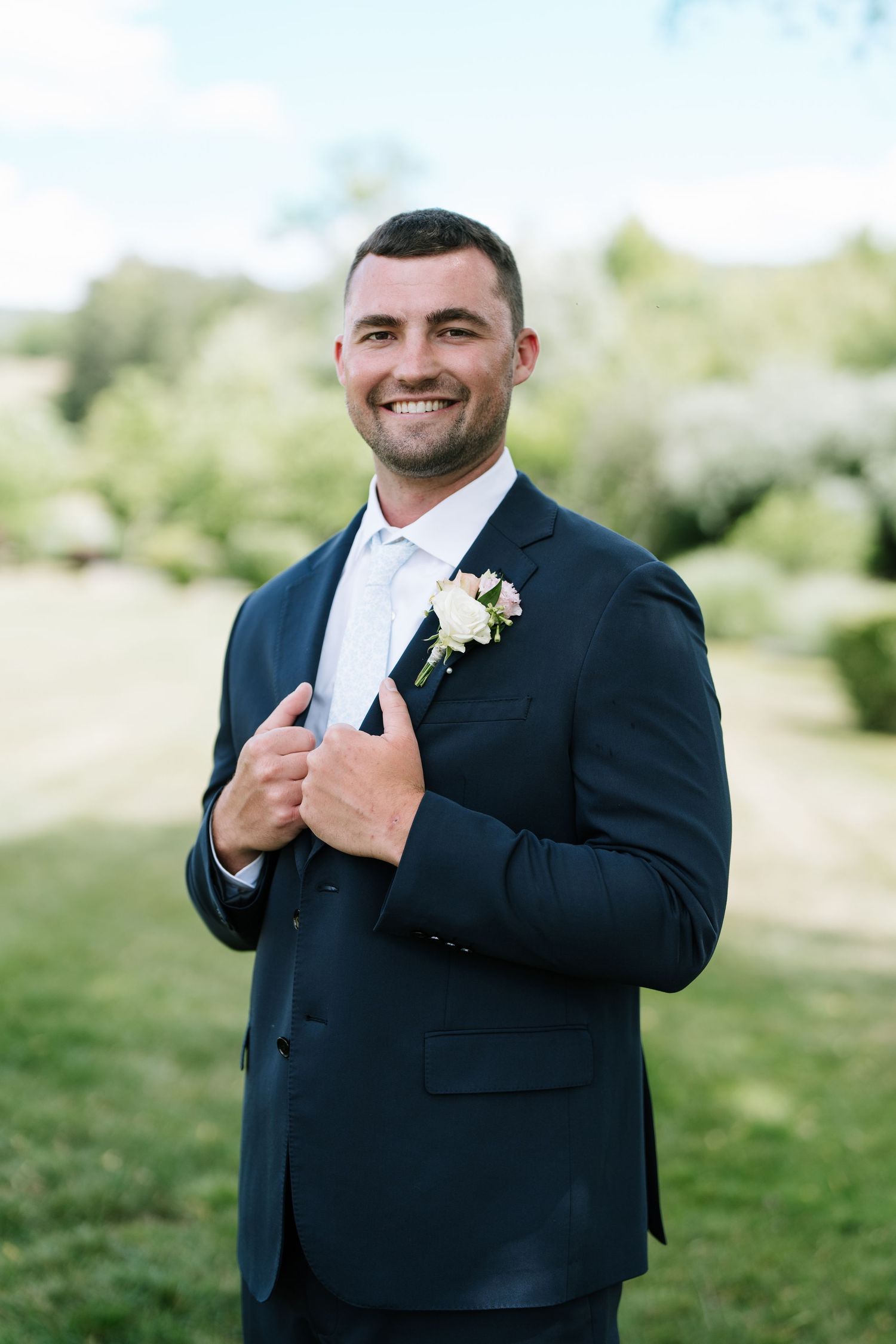 Smiling groom in a navy suit with a white boutonniere posing outdoors on a sunny day.