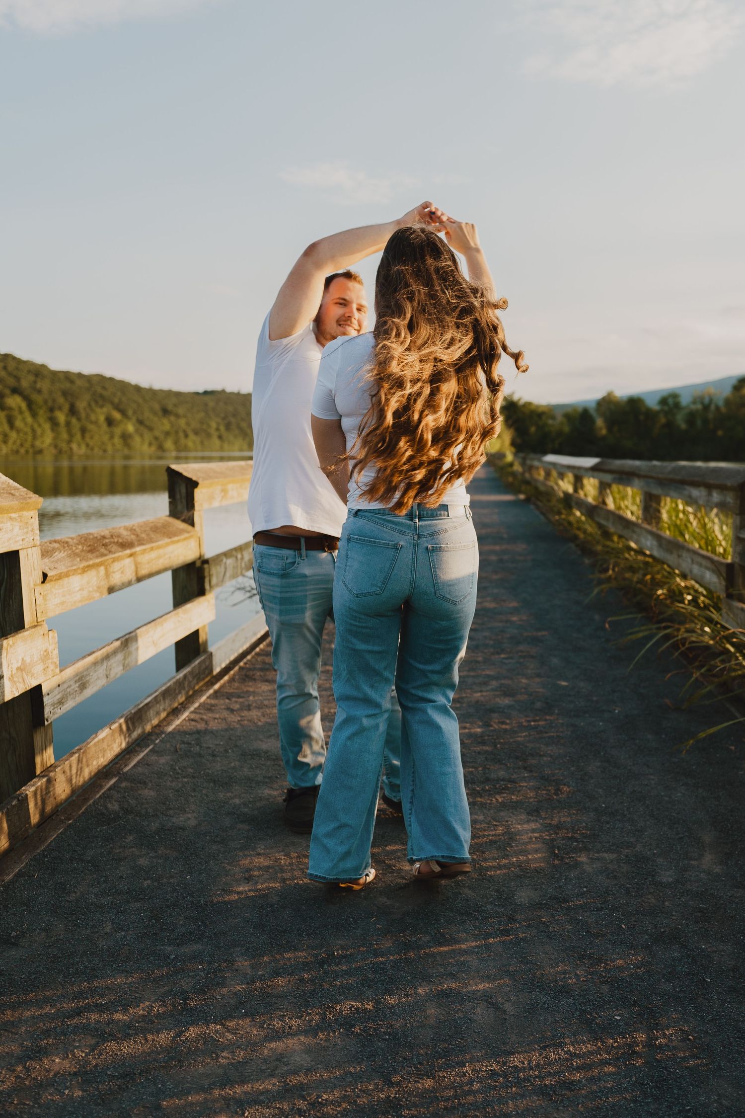 A couple shares an intimate moment along a wooden boardwalk overlooking a tranquil lake at sunset.