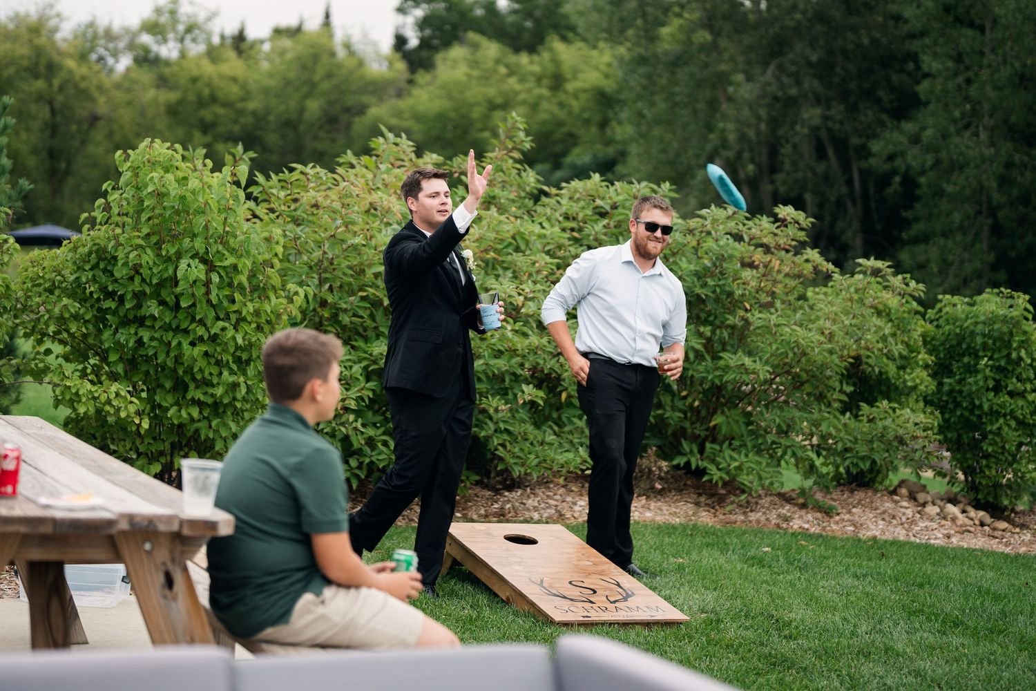 Wedding guests play cornhole lawn game outdoors surrounded by lush green hedges at summer wedding reception.