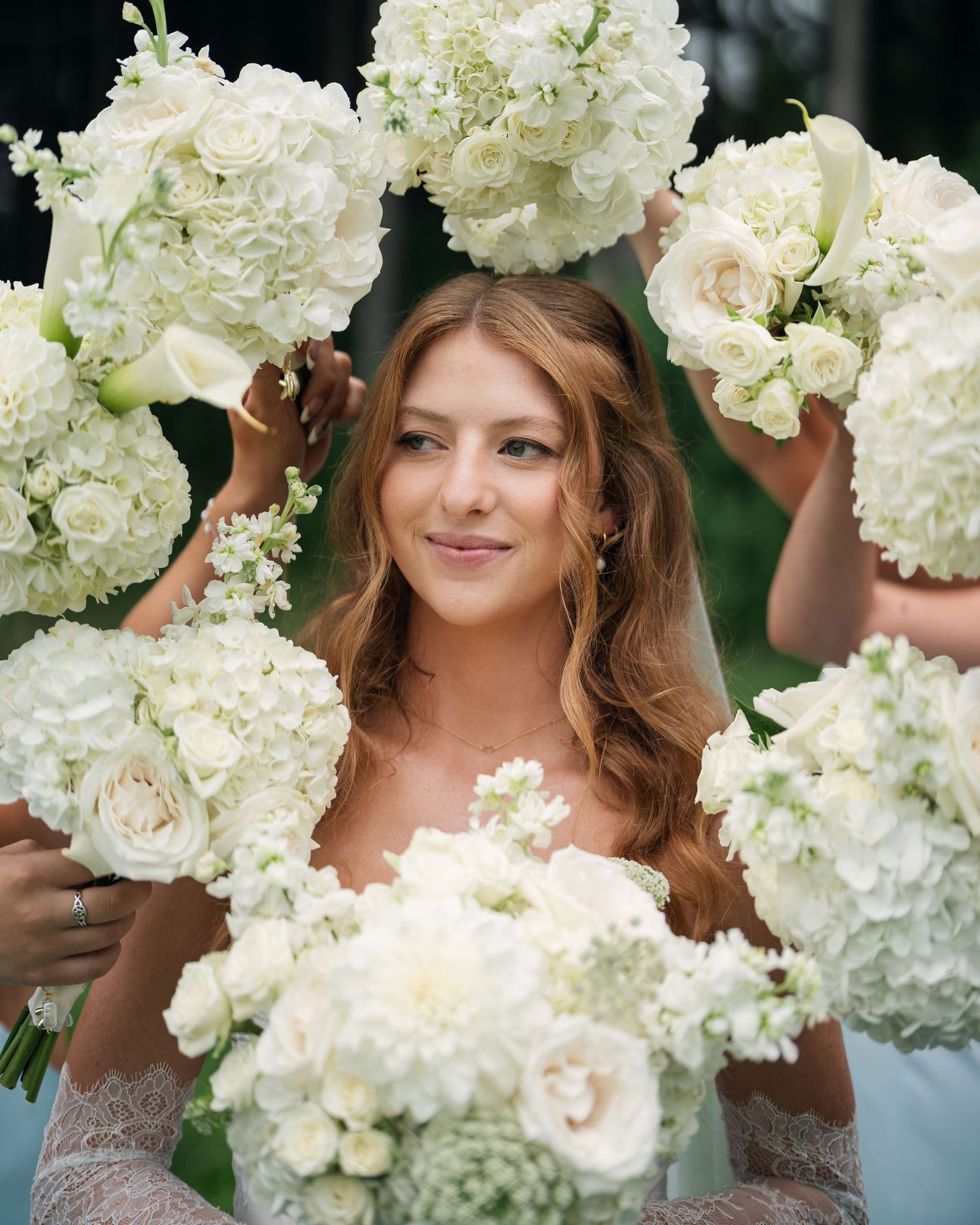 Radiant bride surrounded by lush white rose and hydrangea bouquets held by bridesmaids outdoors.