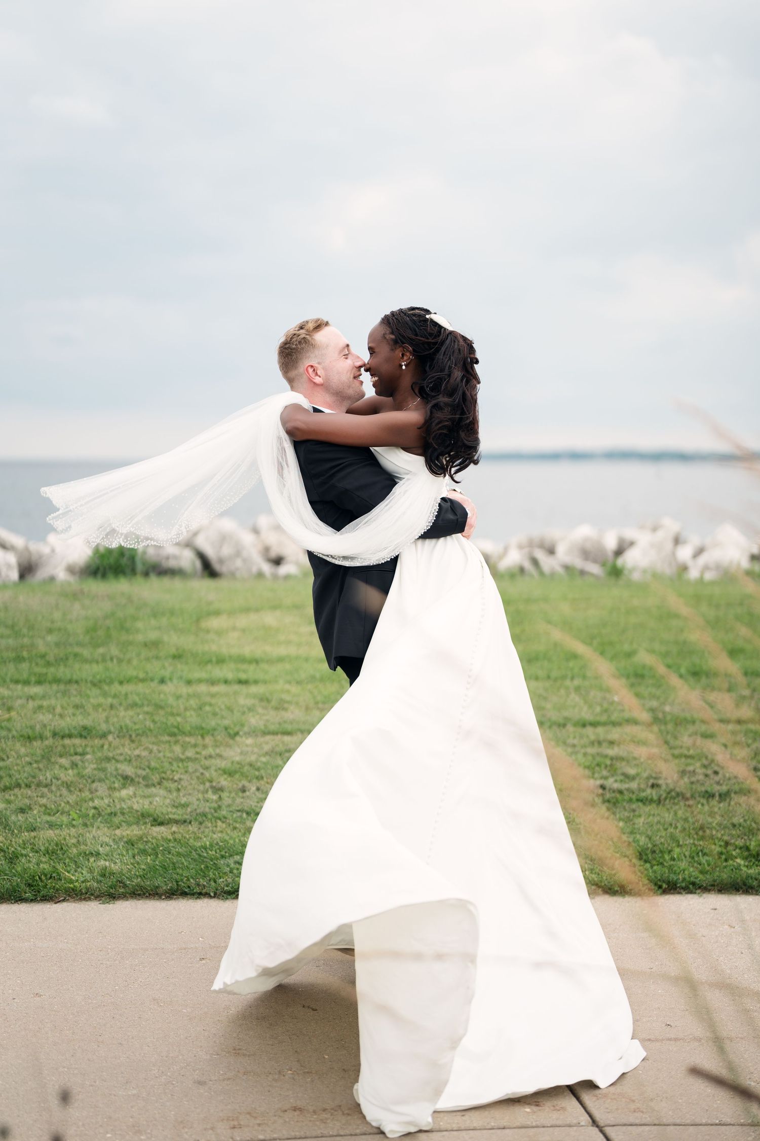 Groom lifts bride in white gown outdoors near waterfront, sharing a romantic moment on their wedding day.