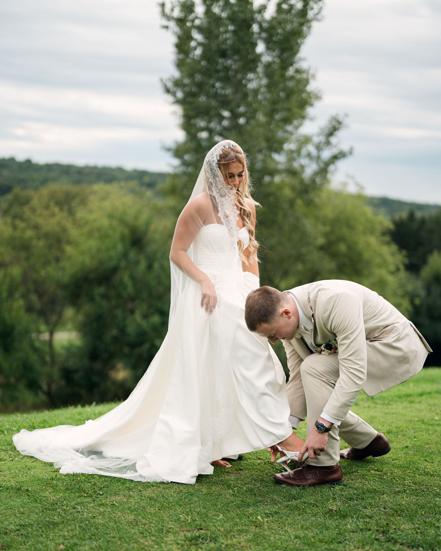 Groom adjusting brides shoes in white wedding gown and veil on green lawn with trees in background.