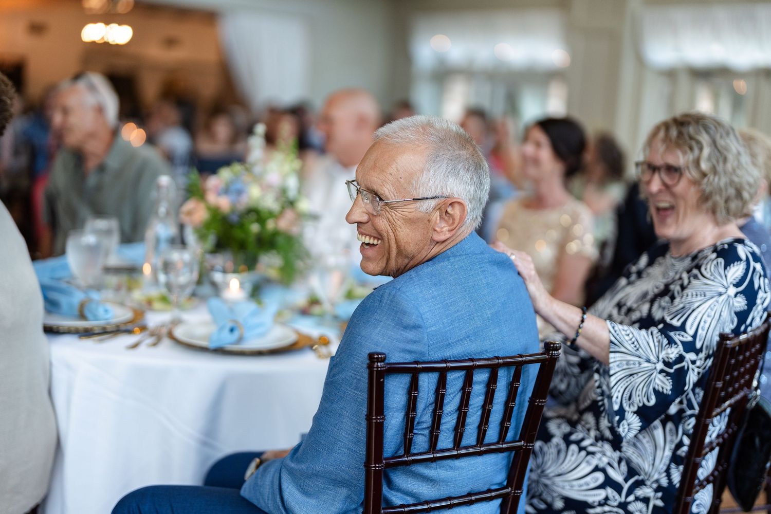 Elderly man in blue blazer and glasses seated at an elegant wedding reception dinner table with guests in background.