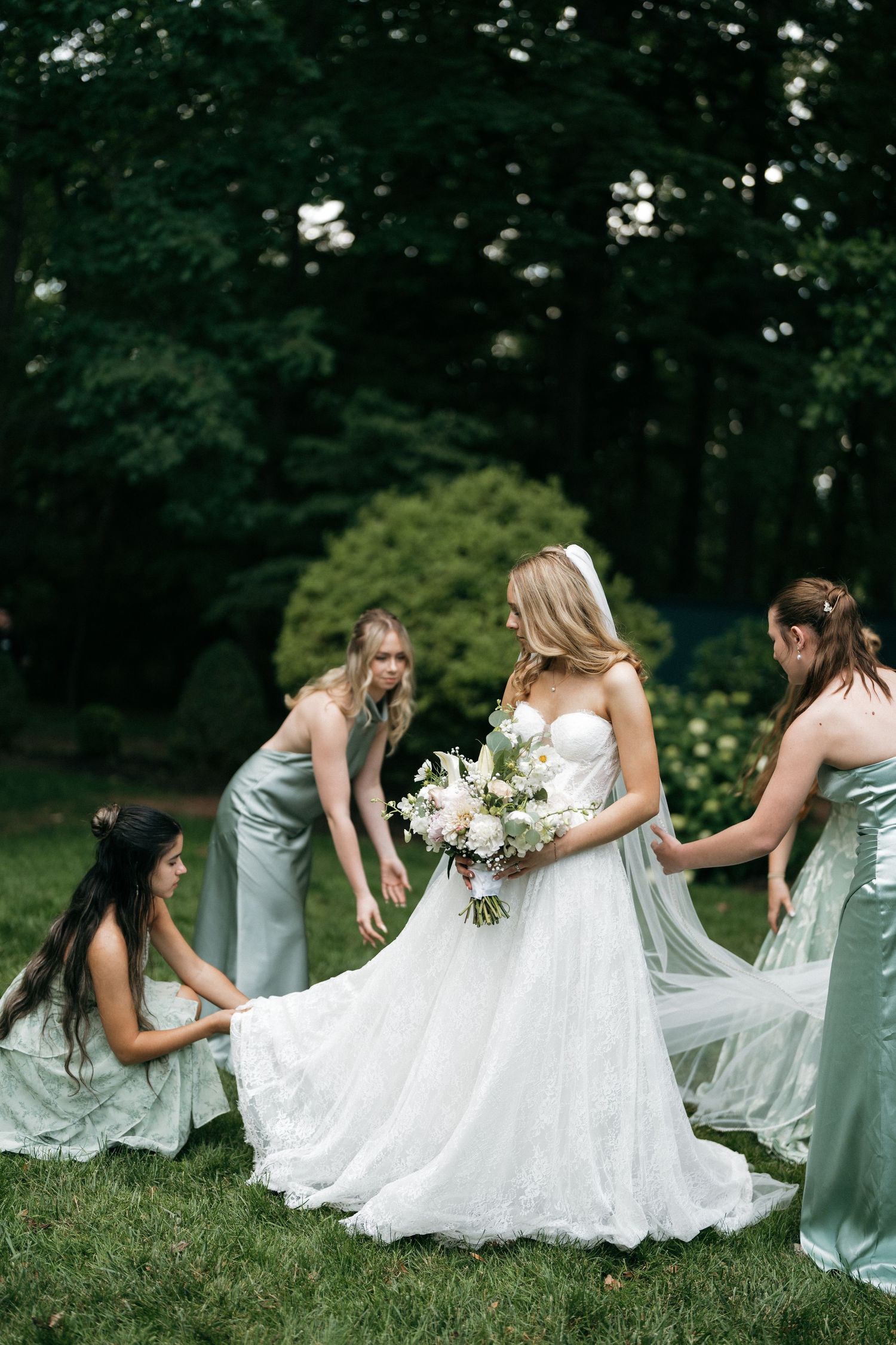Bride in white gown holds bouquet while three bridesmaids in sage green dresses adjust her veil outdoors.