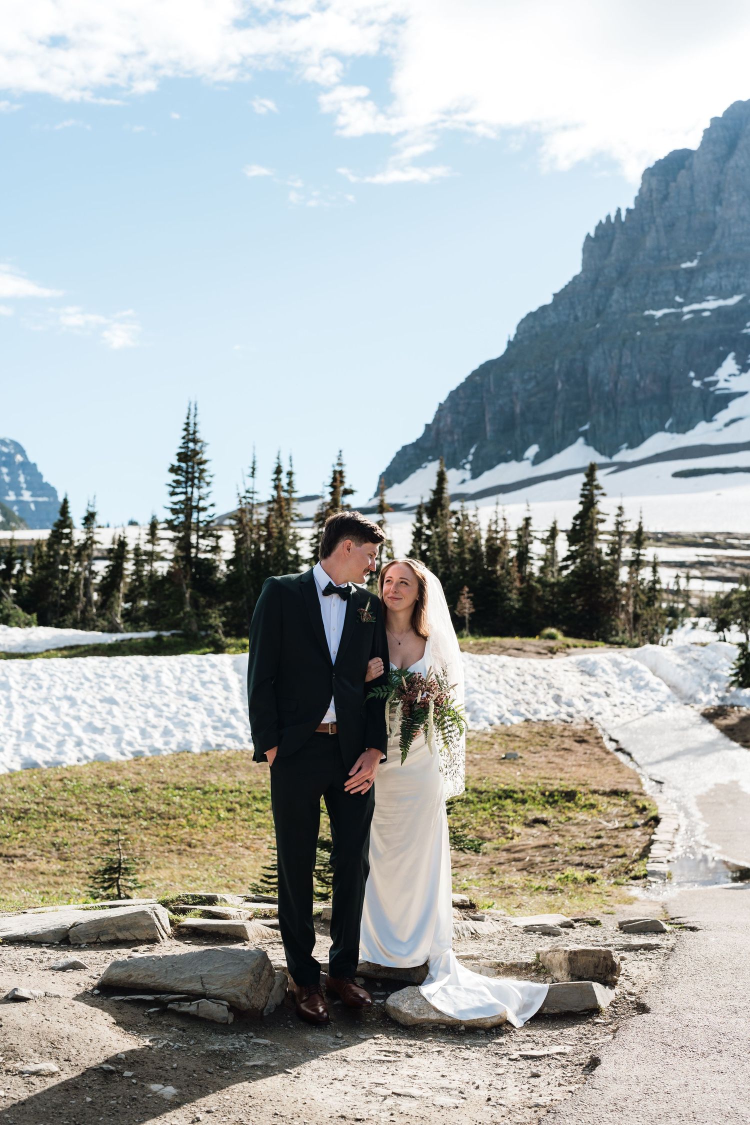 Bride and groom pose in wedding attire with snowy mountain landscape and evergreen trees in background.