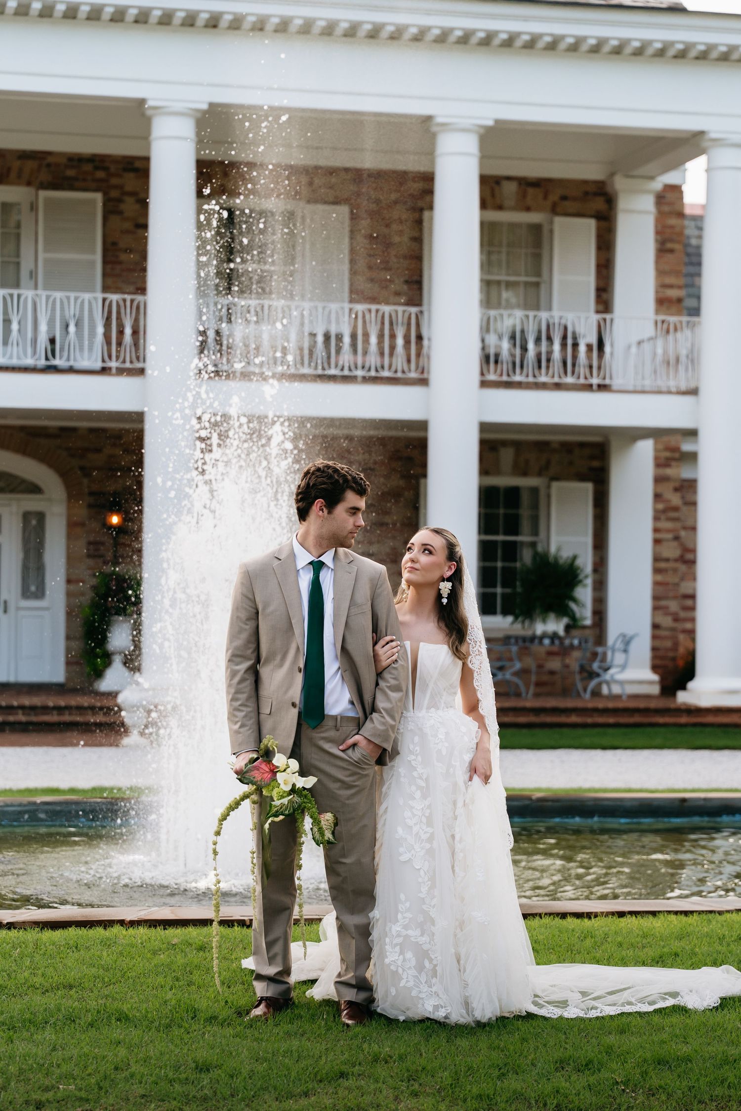 Bride and groom posing in front of a fountain and elegant brick mansion with white columns on their wedding day.
