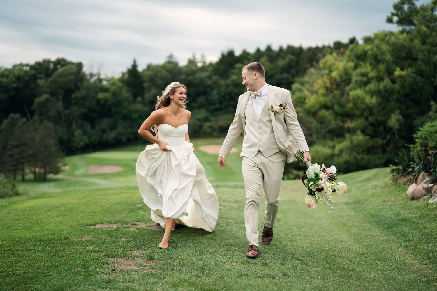 Bride and groom joyfully running on lush green golf course, bride in white gown, groom in beige suit holding bouquet.