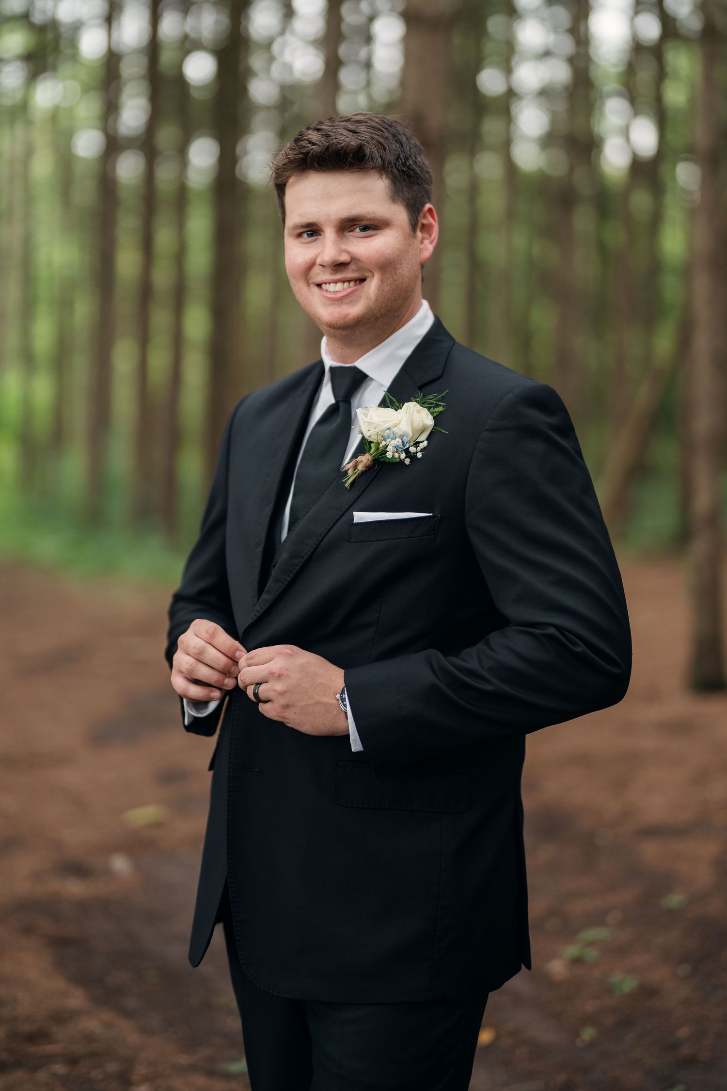 Handsome groom in black tuxedo with floral boutonniere smiles confidently in a pine forest setting.