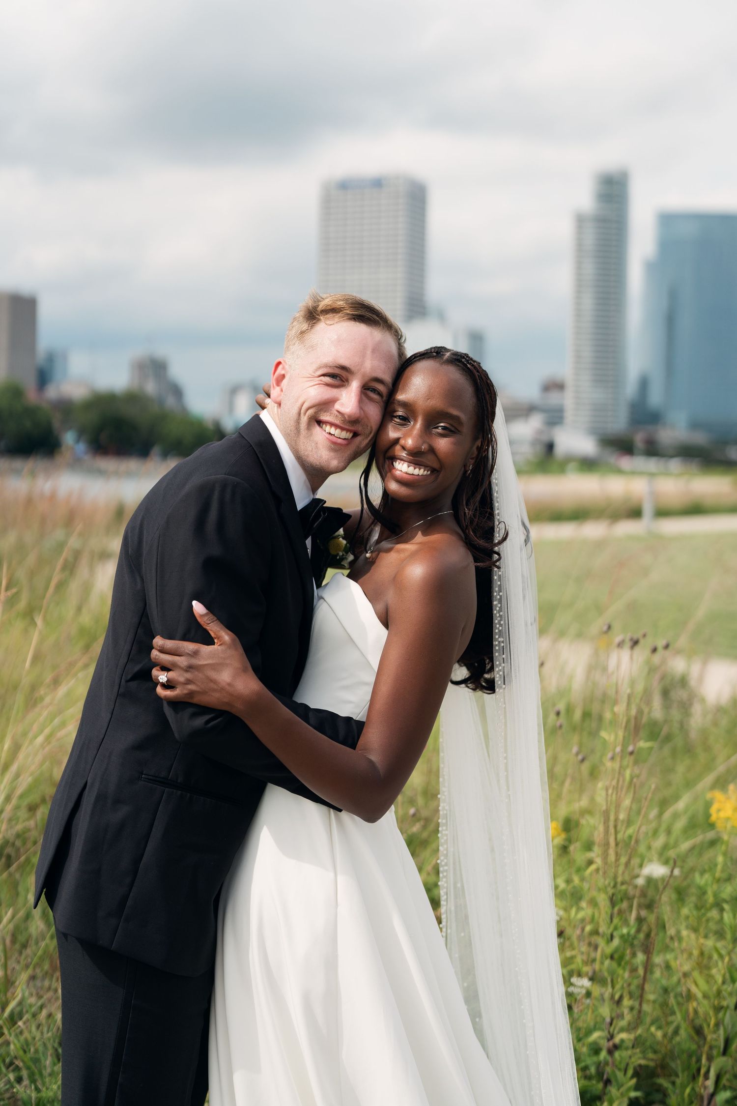 Smiling interracial bride and groom embracing in a field with a city skyline in the background.