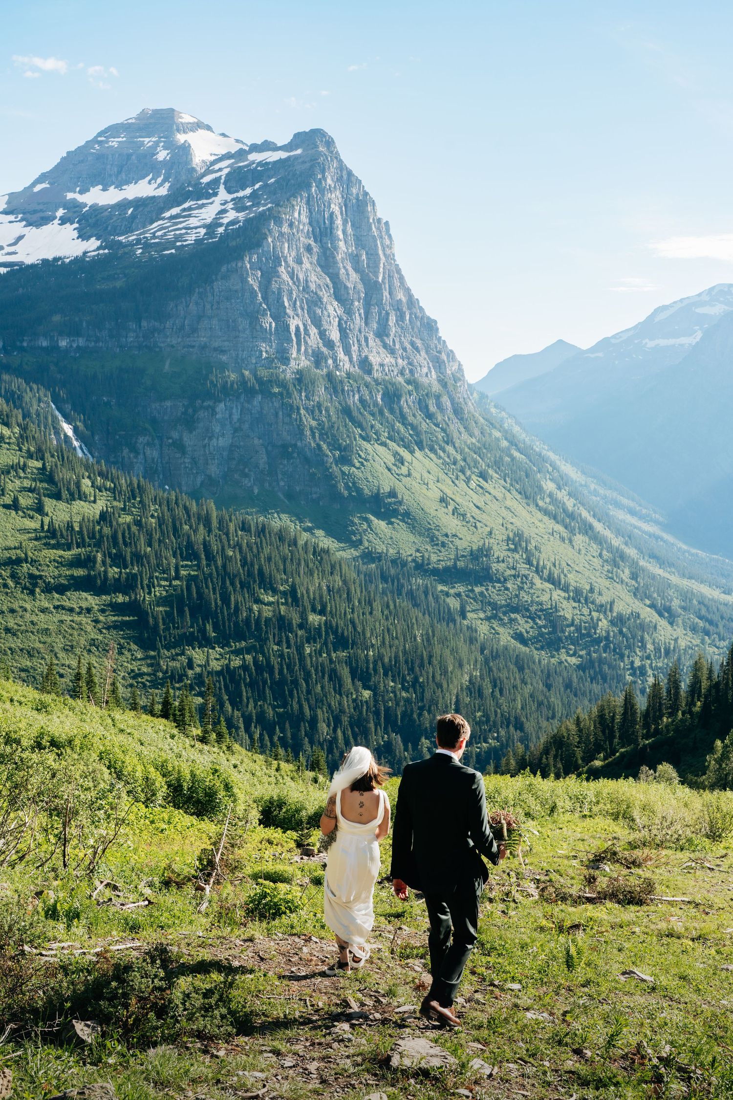 Bride and groom walk hand-in-hand on a mountain trail with stunning green valleys and snow-capped peaks in the background.