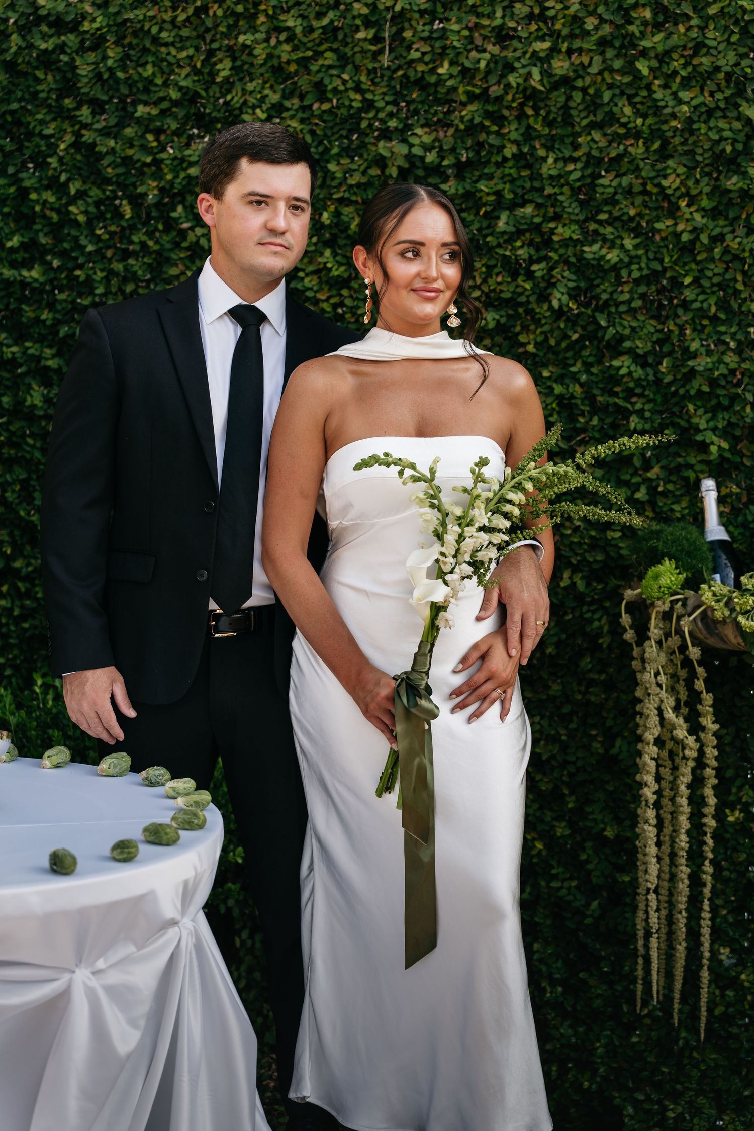 A couple in formal wedding attire poses against a lush green hedge wall at an elegant outdoor venue.