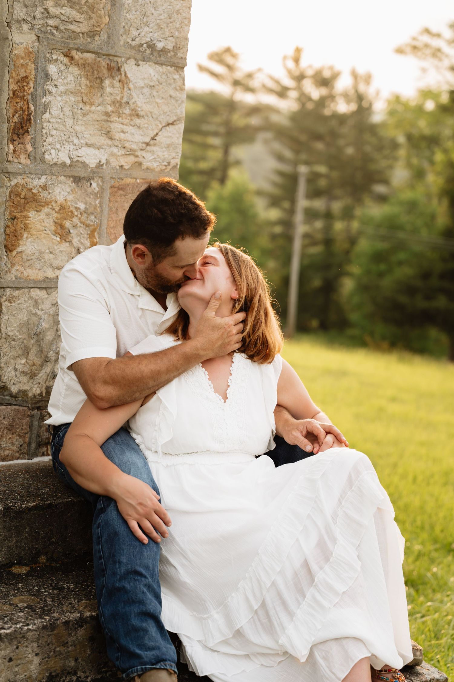 A romantic couple sharing an intimate moment while sitting against a stone wall at sunset in a grassy field.