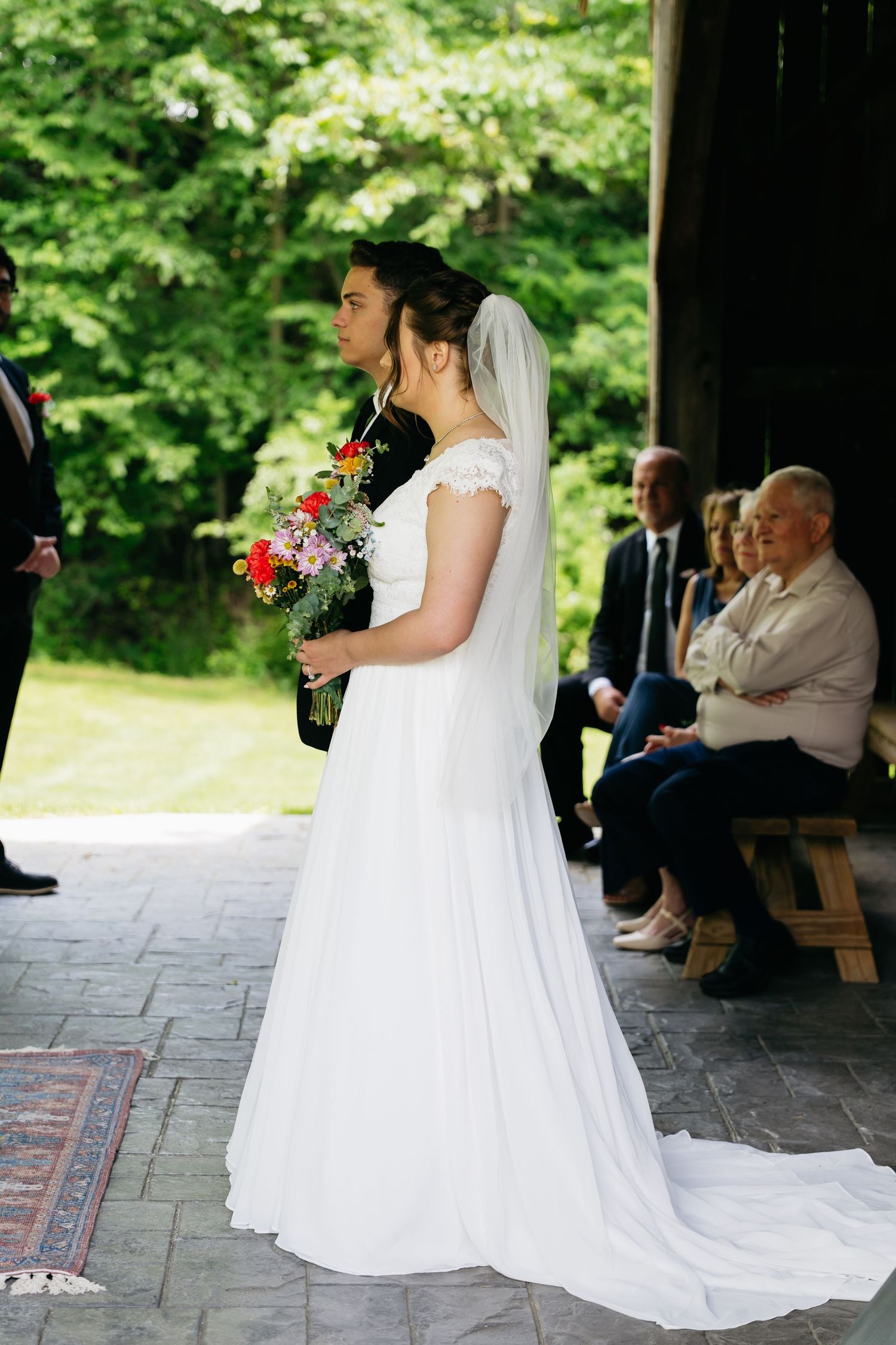 A bride in a flowing white wedding dress with cap sleeves and long veil holds a colorful bouquet during an outdoor ceremony.