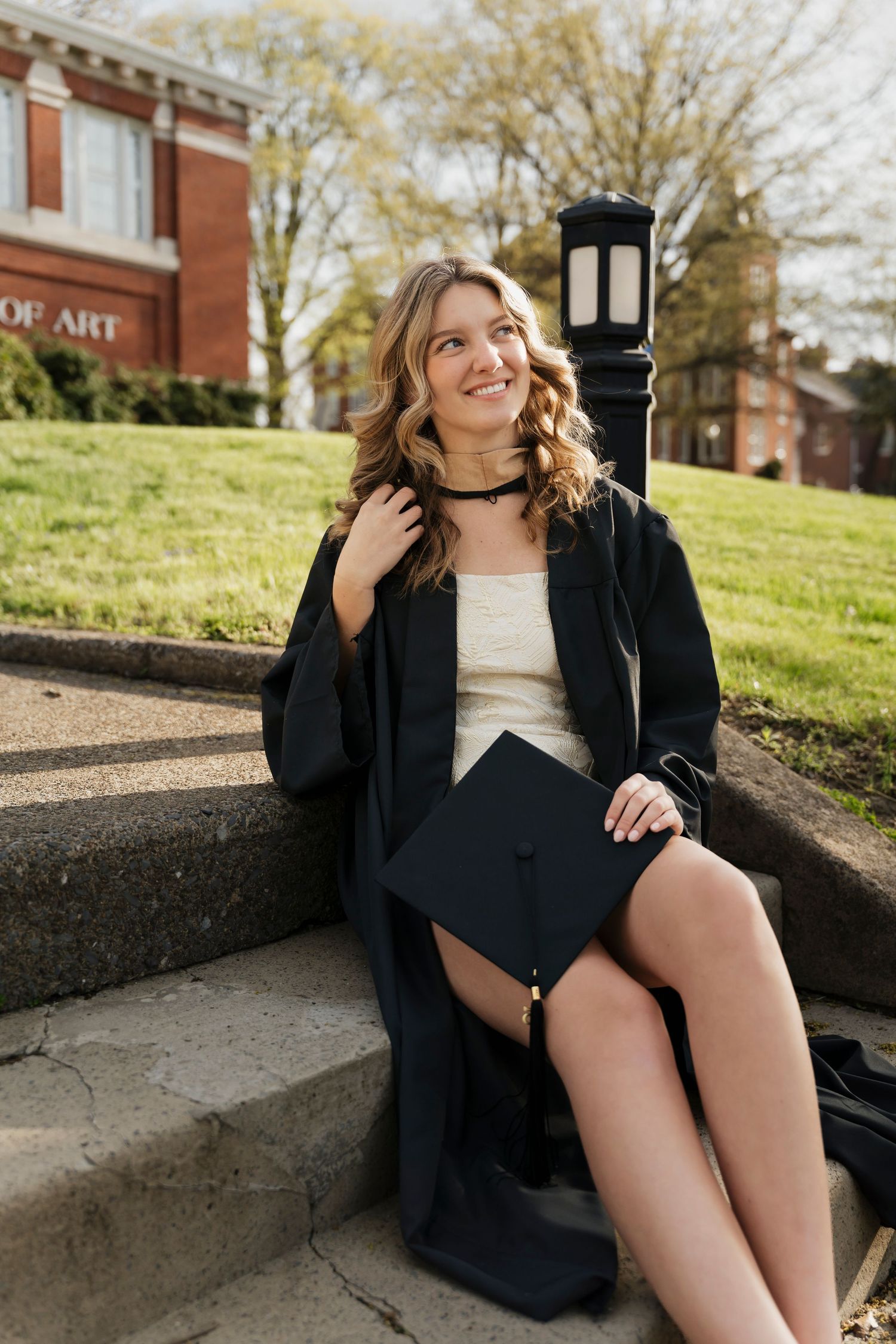 Graduate in traditional black gown sits on concrete steps during an outdoor spring photoshoot.