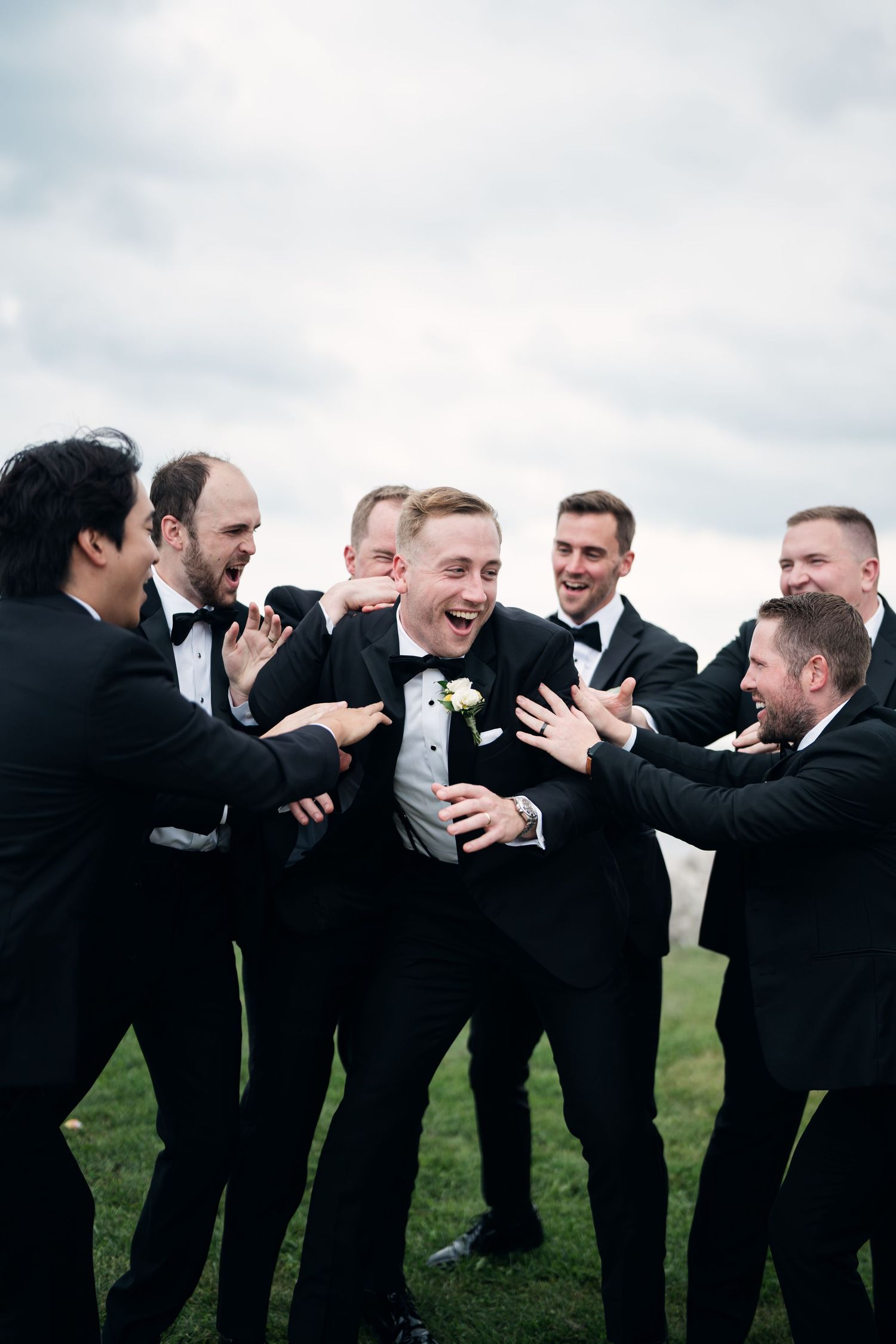 Groom laughing with groomsmen in black tuxedos outdoors on green grass under cloudy sky at wedding.