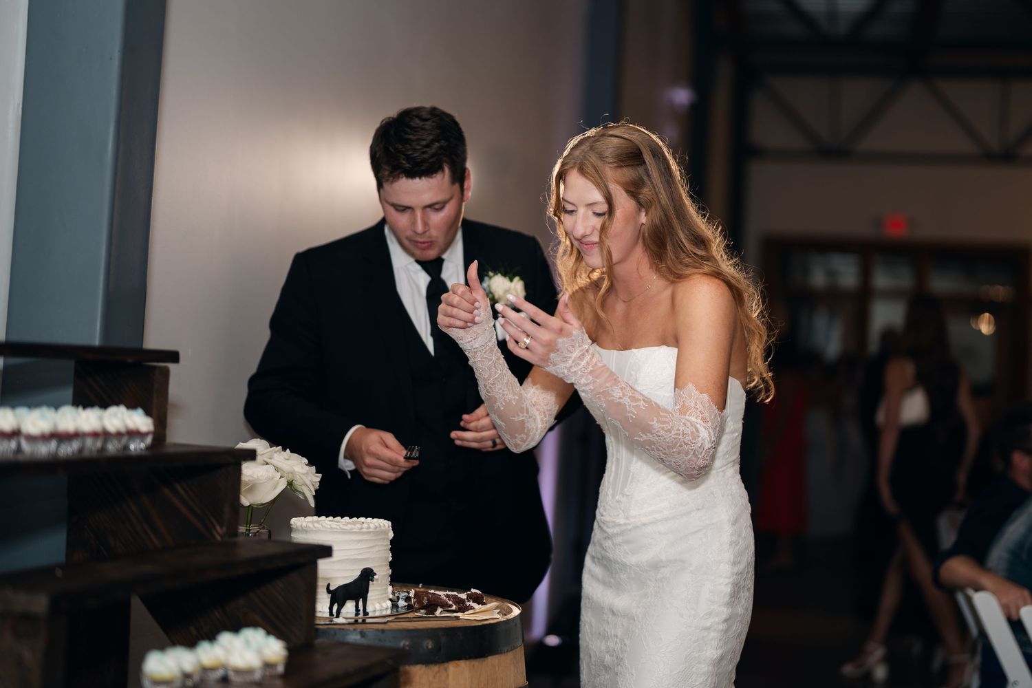 Bride in lace gown and groom in black tux cut wedding cake together at elegant indoor reception.