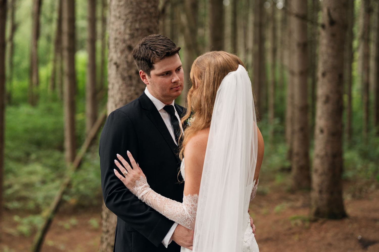 Bride and groom share an intimate moment in a lush forest, bride wearing white gown and veil, groom in black suit.