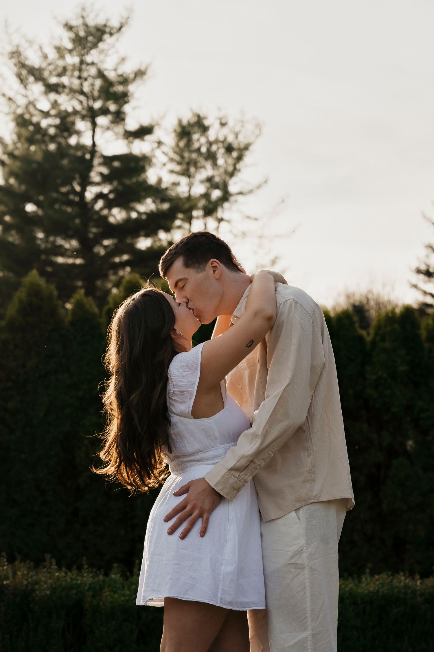 A couple embraces tenderly in a romantic outdoor setting with evergreen trees silhouetted against the sunset.
