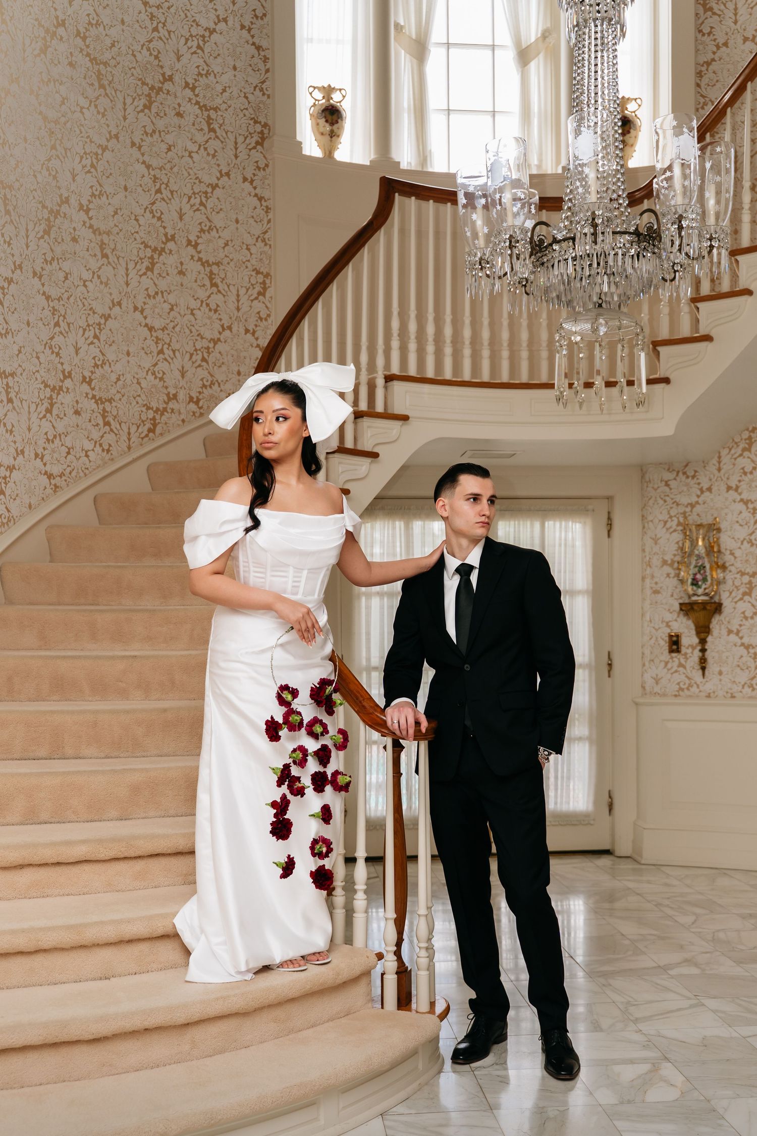 Elegant bride in white off-shoulder gown with bow headpiece holds red bouquet beside groom in black suit on grand staircase.