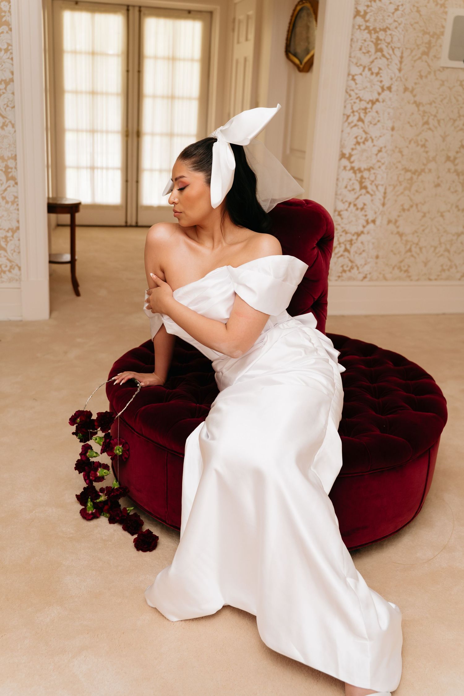 Bride in white off-shoulder gown with bow headpiece, seated on red velvet chaise, holding dark roses.