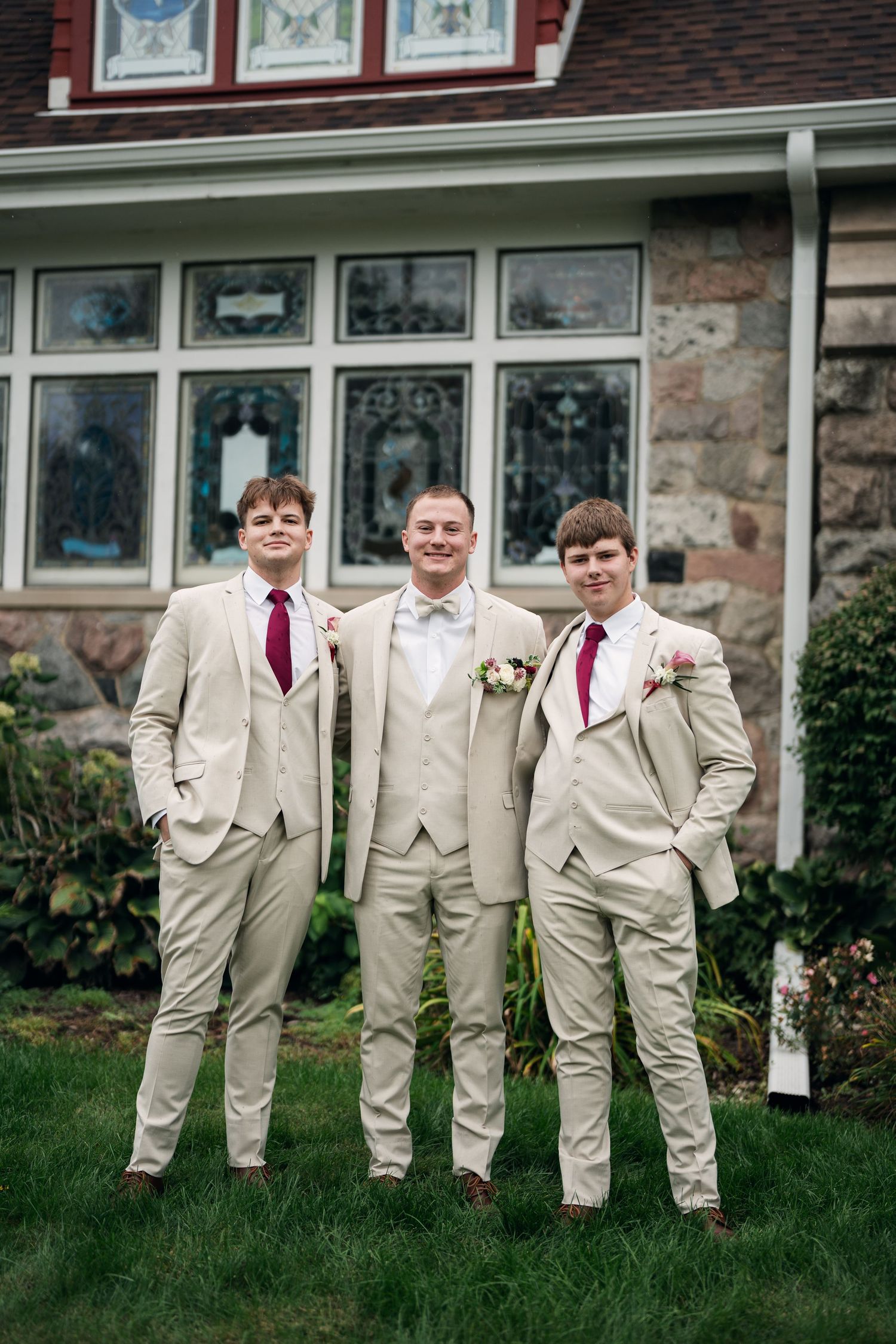 Groom and two groomsmen in tan suits with burgundy ties pose outside a stone building with stained glass windows.