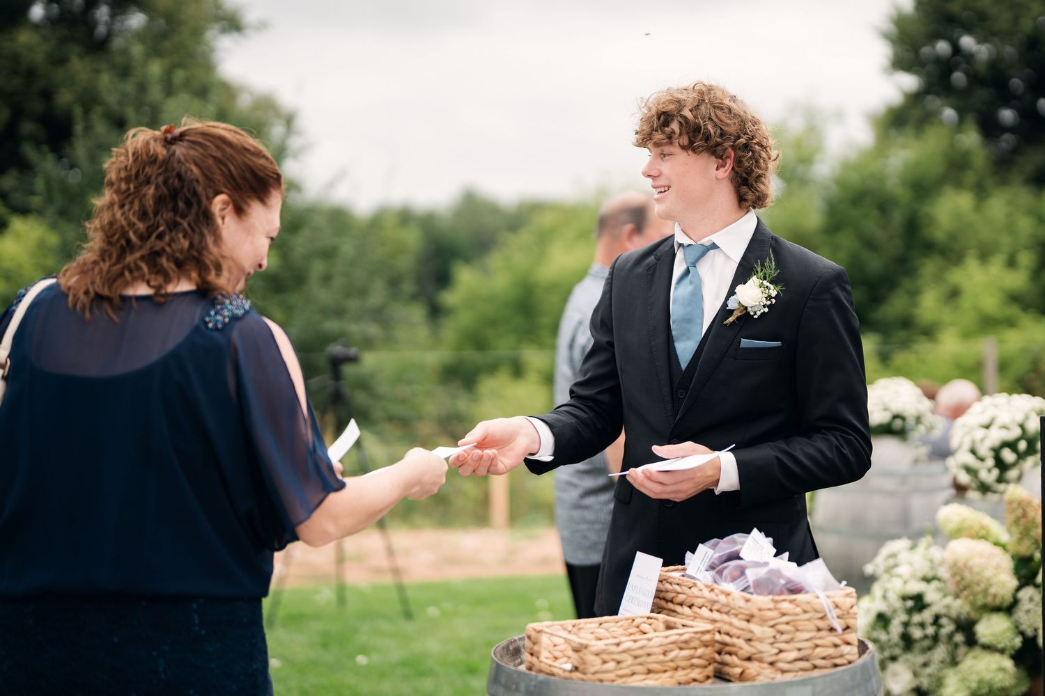 Young groom in black suit with teal tie handing program to guest at outdoor wedding ceremony with floral decorations.