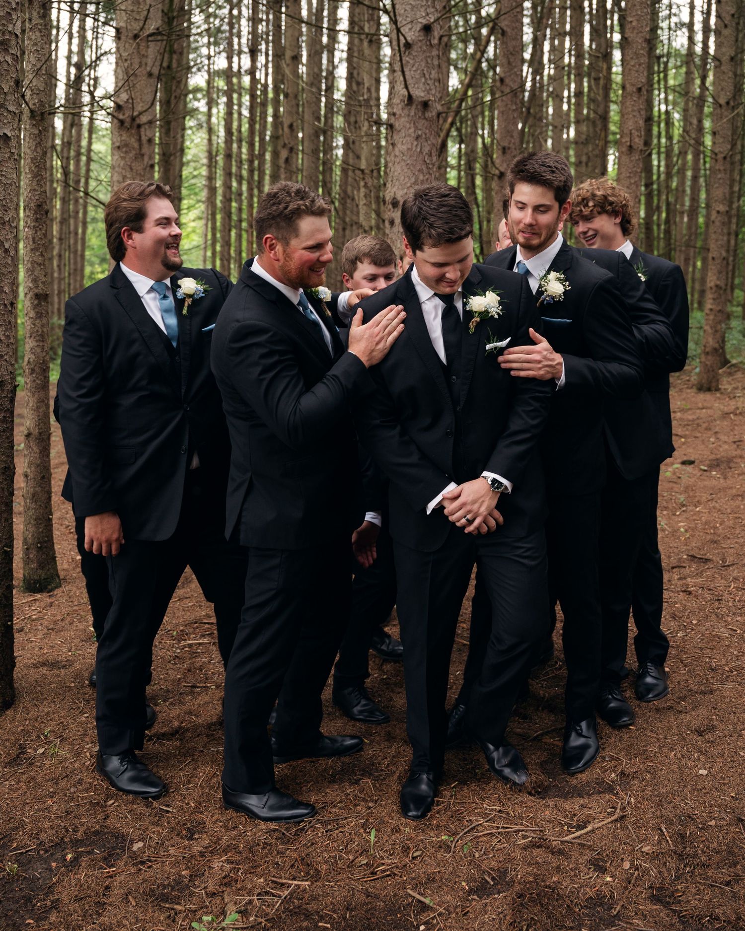 Groomsmen in black suits laughing together in a pine forest during a wedding photo session.