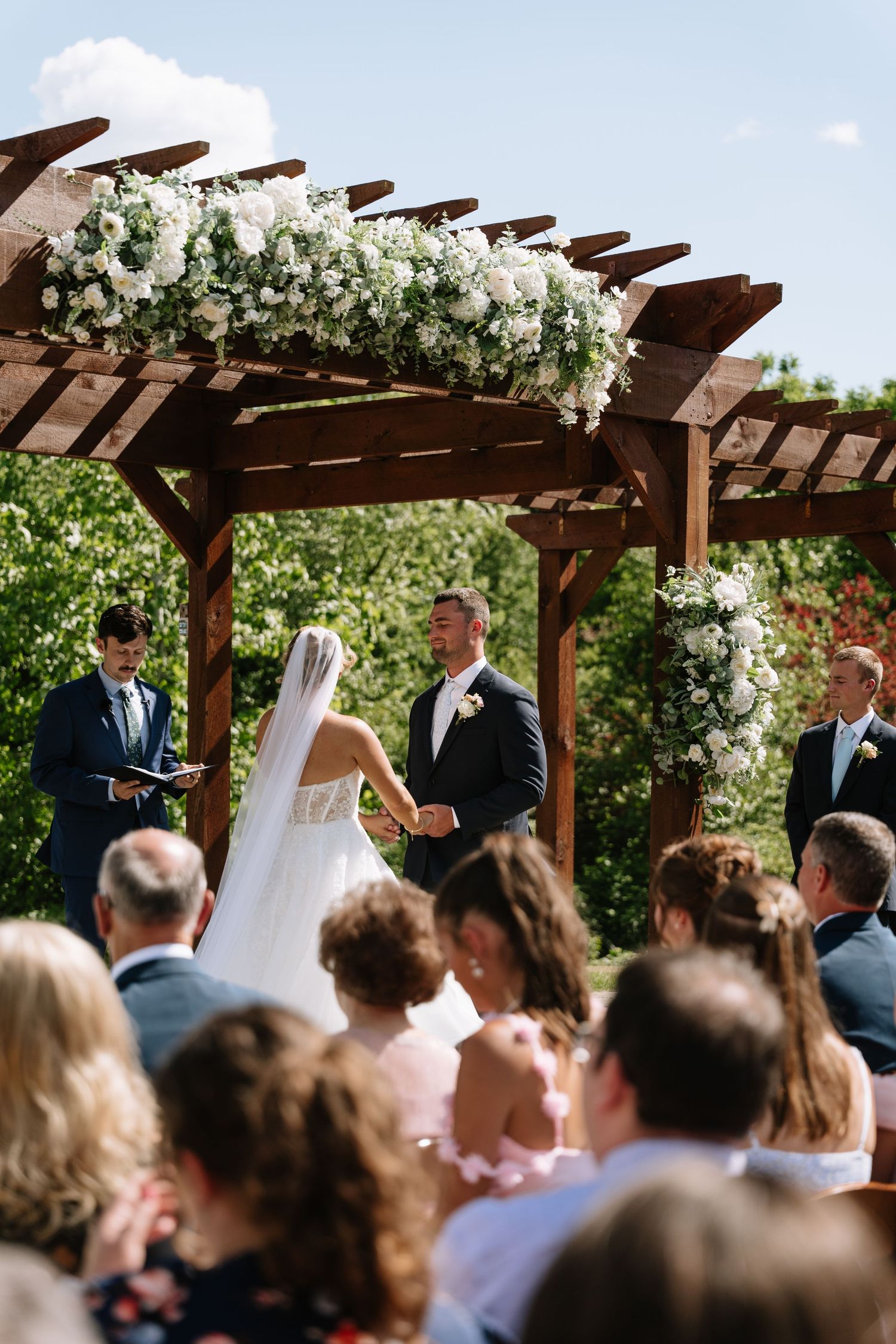Outdoor wedding ceremony under wooden pergola with white flowers, couple exchanging vows before guests.