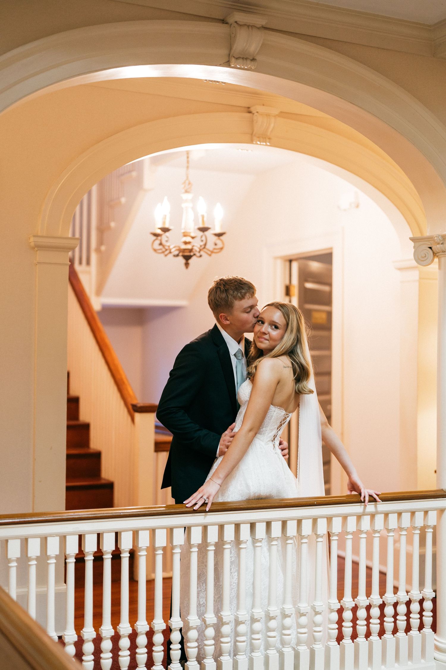 Groom kissing smiling bride beside white banister under chandelier in elegant staircase of historic venue.