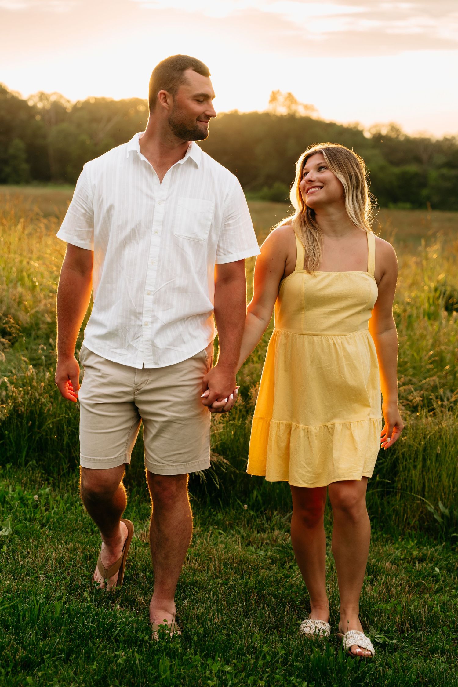 Couple holding hands walking through a golden meadow at sunset wearing yellow dress and white shirt.
