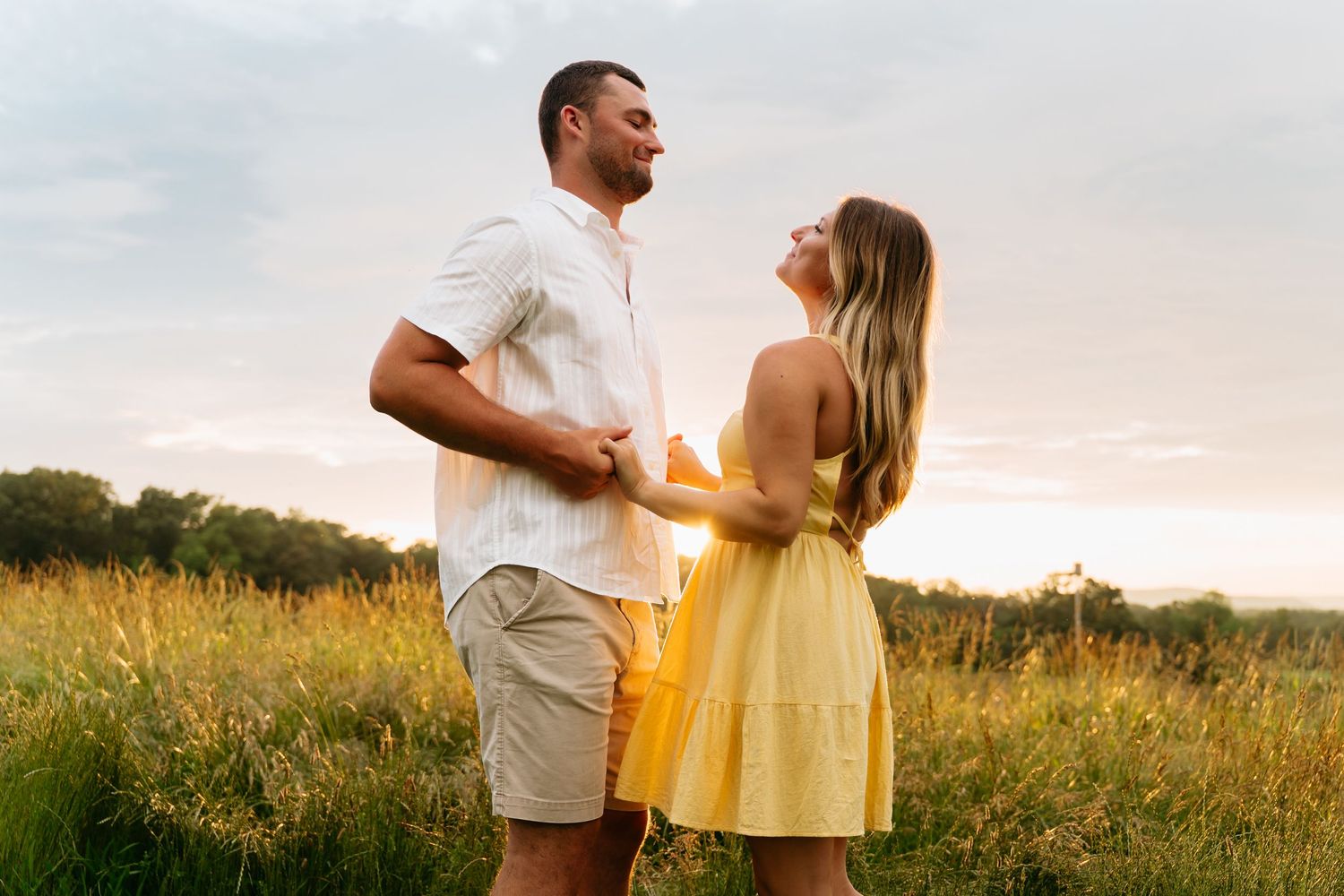 A couple in summer attire embraces while standing in a golden meadow at sunset.