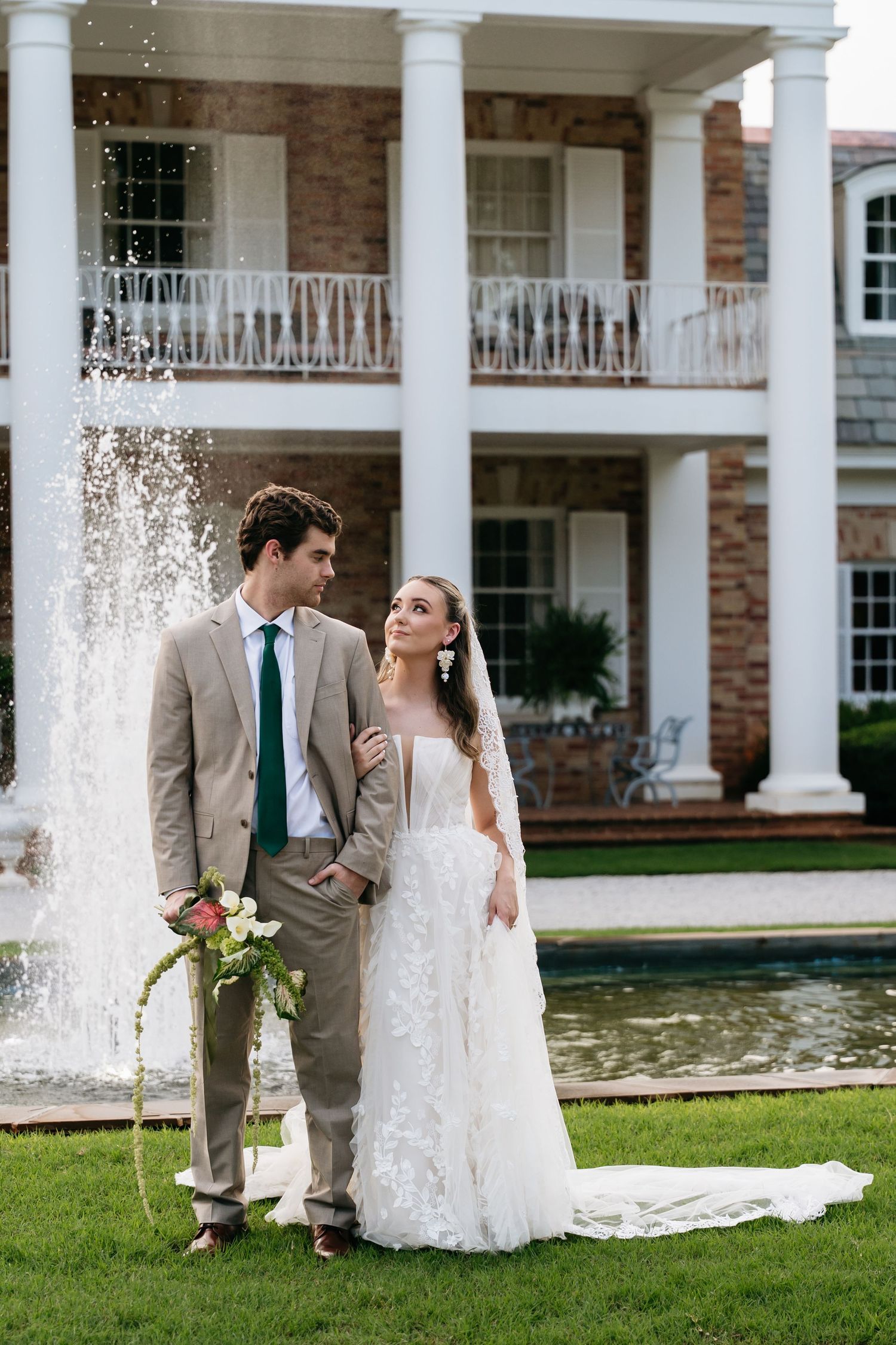 A bride and groom share romantic moments in front of a stately mansion with white columns and a reflecting pool.
