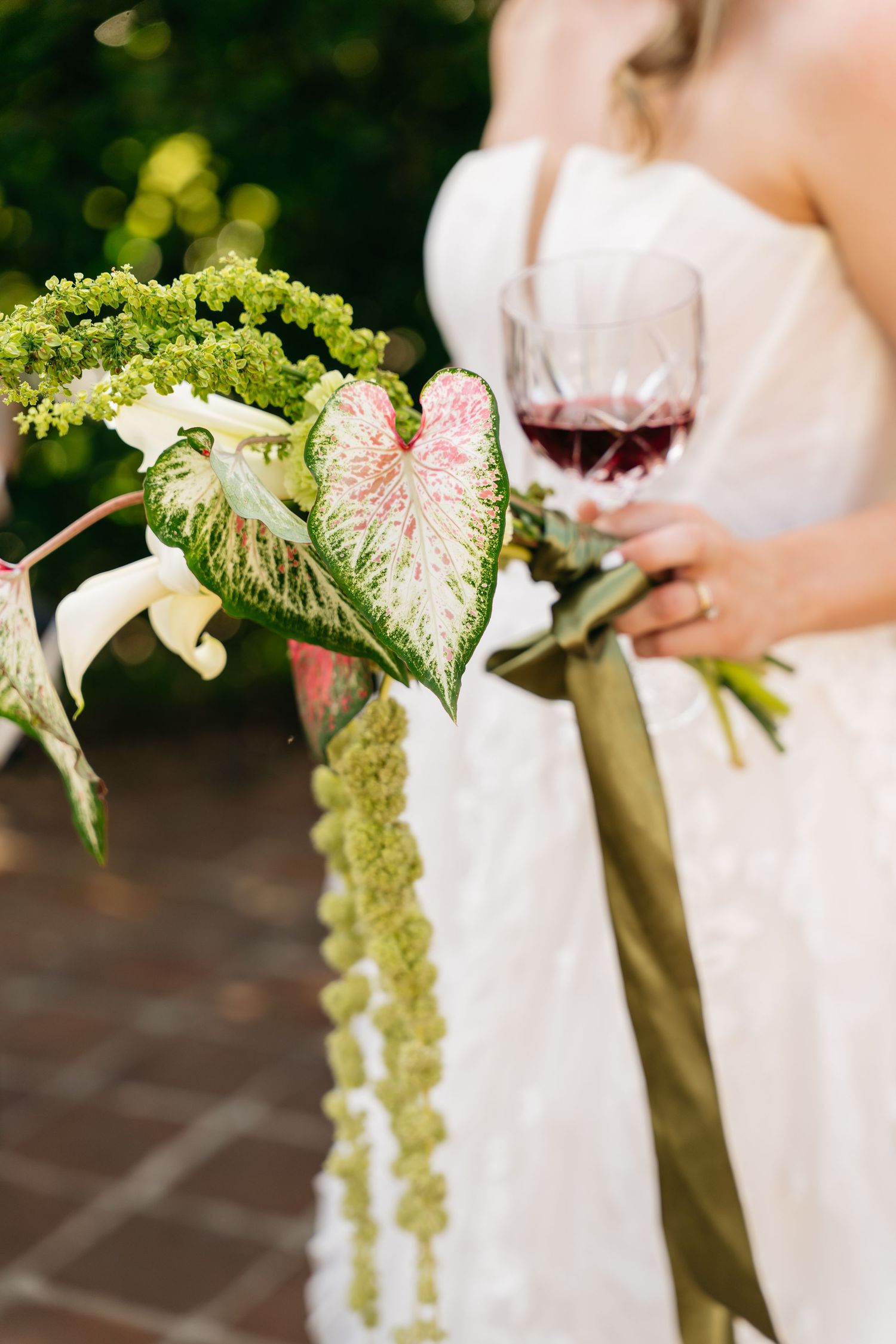 Close-up details of white wedding dress with green and pink floral bouquet accents and red wine glass.