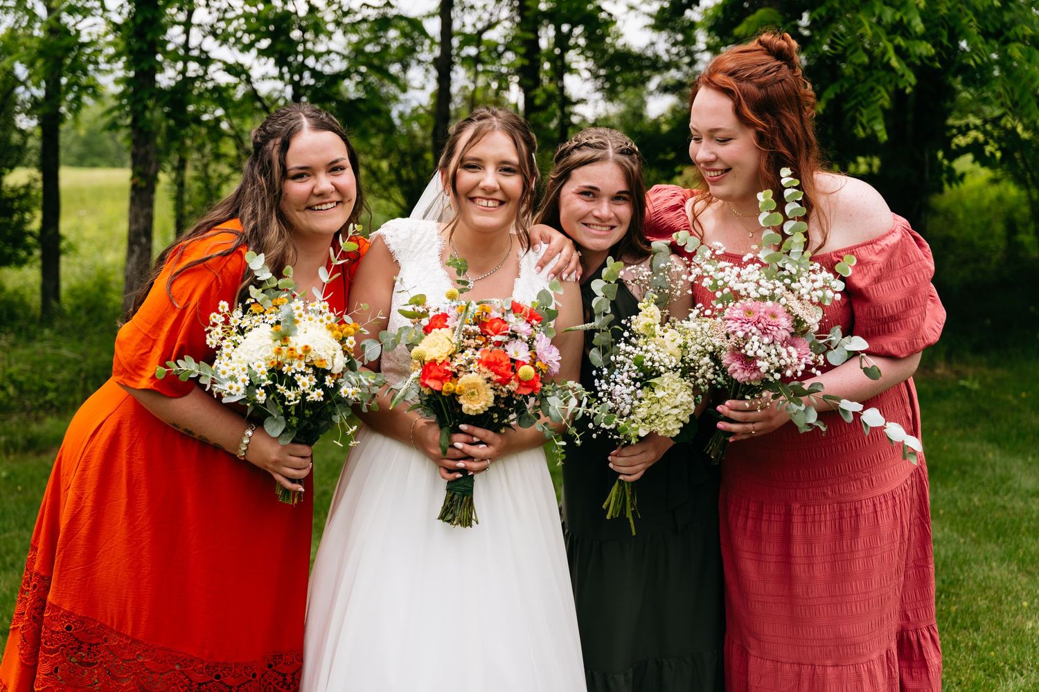 Bride and bridesmaids hold vibrant floral bouquets while wearing dresses in white, red, and dark colors on a grassy lawn.